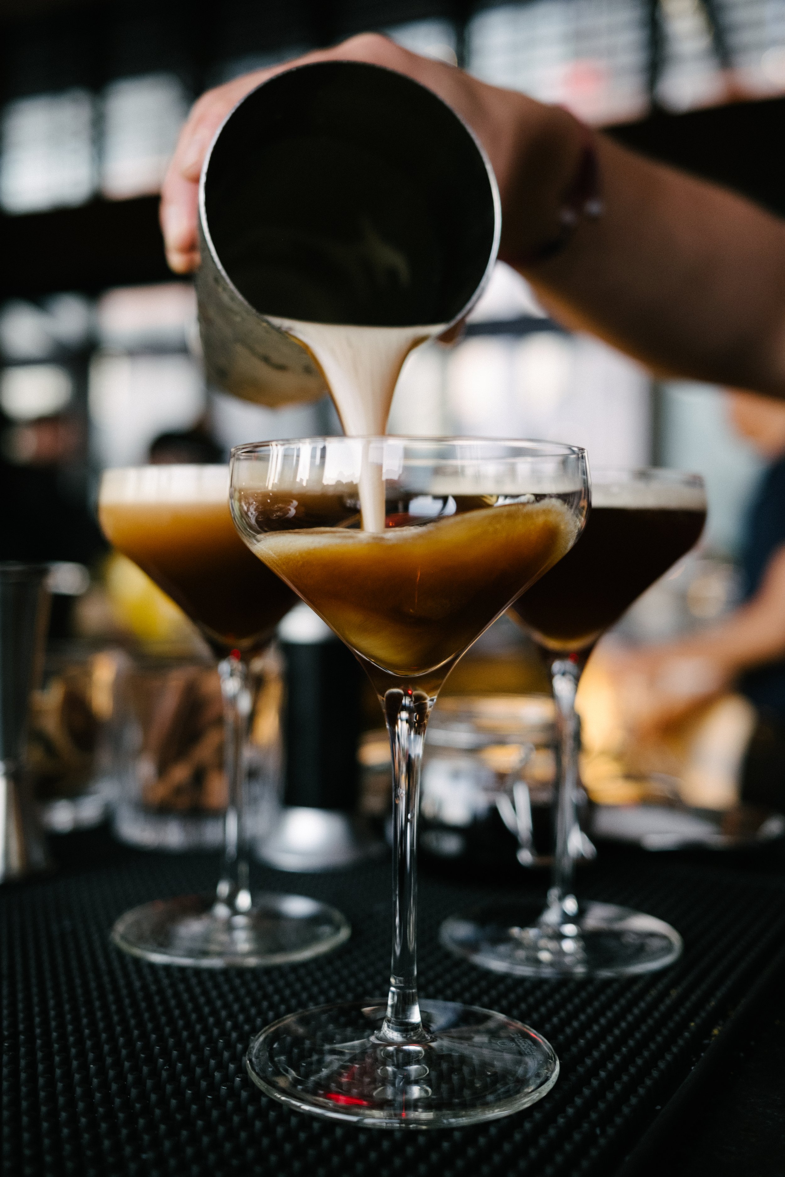 A bartender pouring a creamy liquid into a cocktail glass, with other cocktails visible in the background.