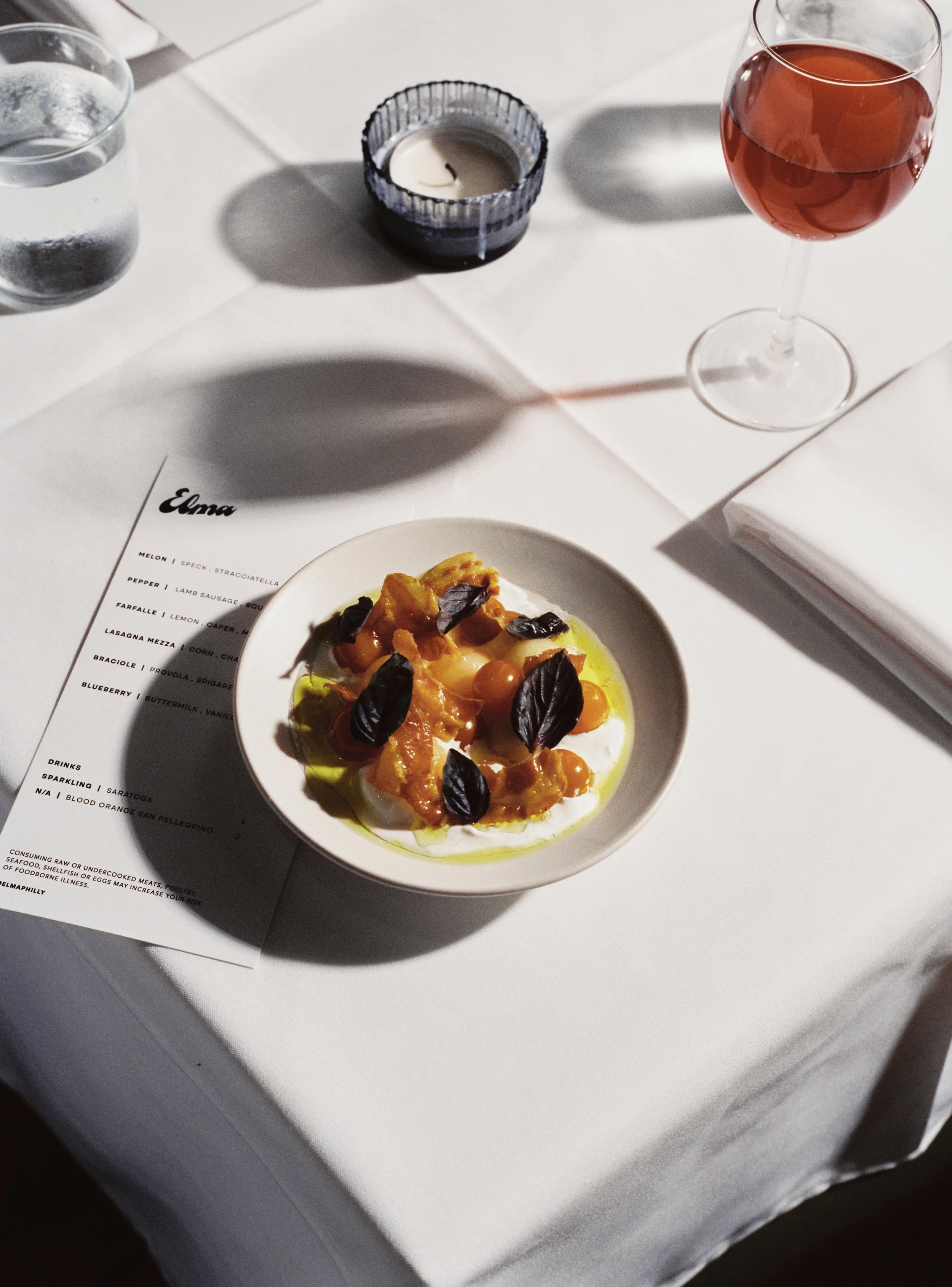 A white plate of burrata cheese topped with yellow and orange fruits and black basil leaves, garnished with olive oil, on a white table with a menu partially visible and a glass of red wine.