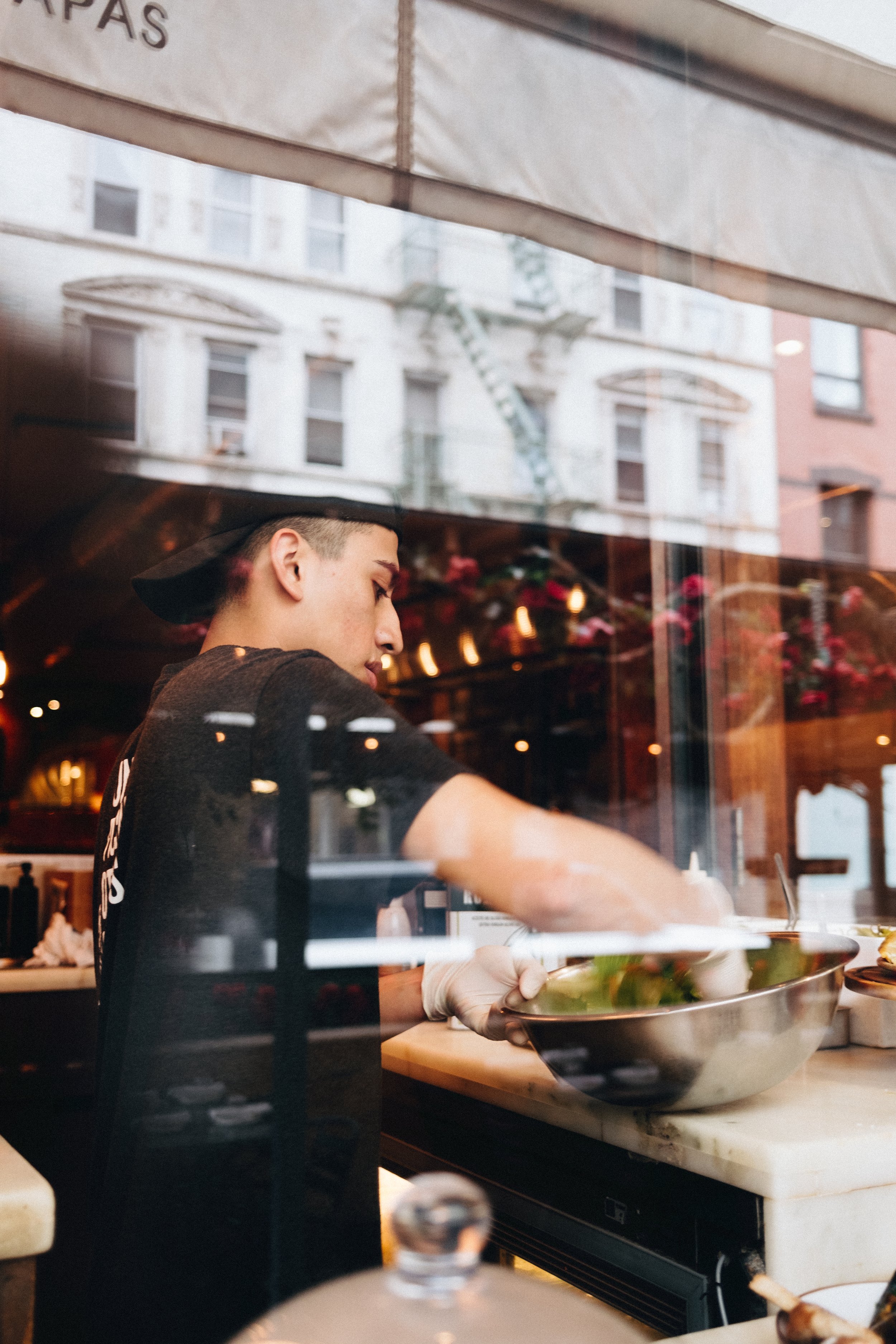 A young man wearing a black cap and black shirt preparing food in a restaurant kitchen, seen through a glass window with reflections of buildings and street outside.