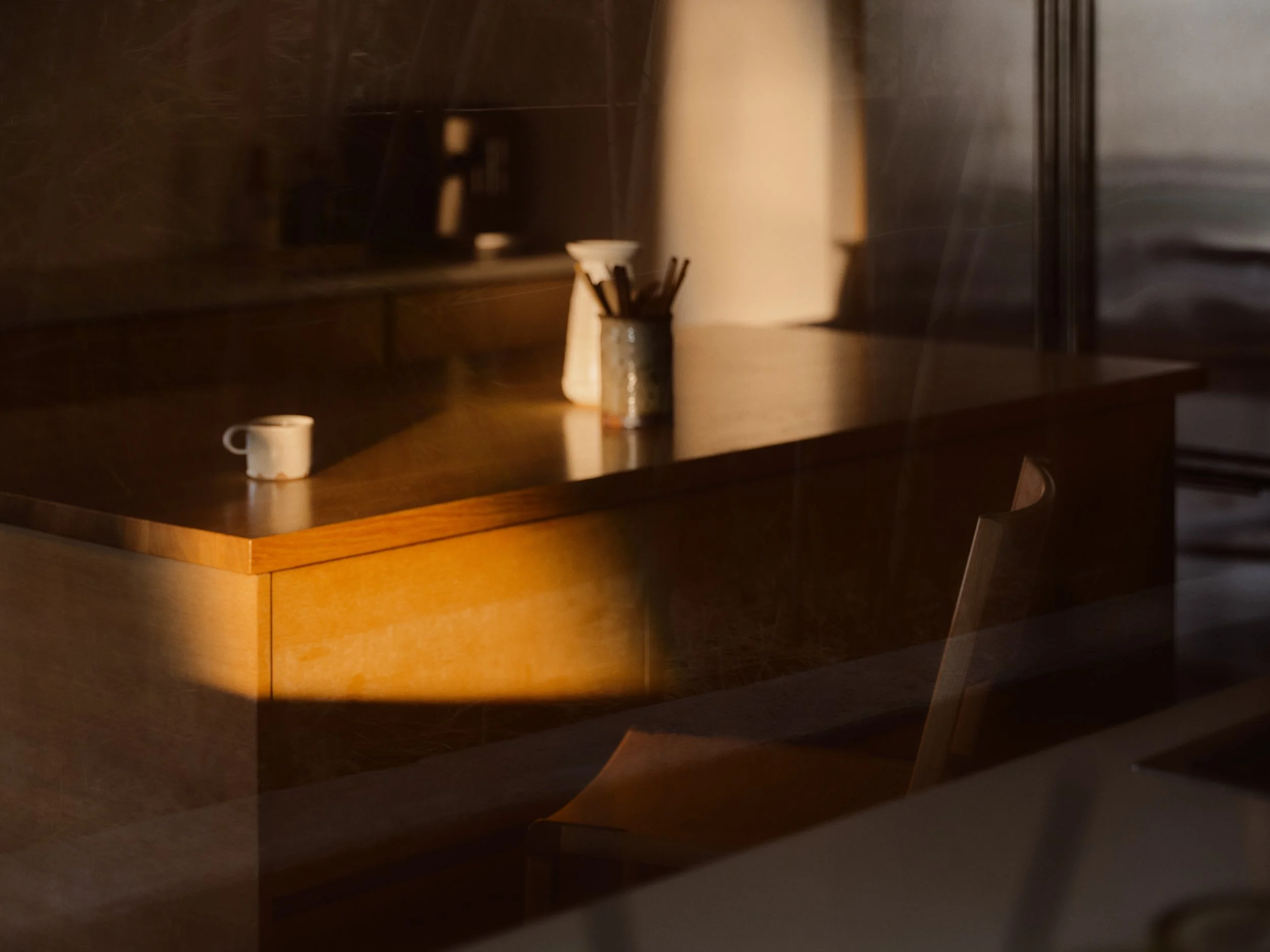 A wooden table with a white coffee mug, a glass jar with utensils, a white tea light holder, and a white lamp in a dimly lit room.