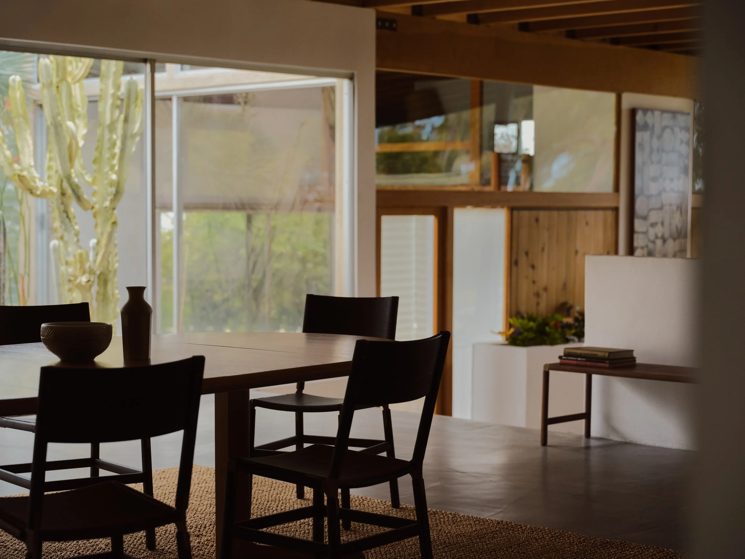 A living room with large windows, wooden beams on the ceiling, a dining table with chairs, decorative vases, and a stone wall section visible in the background.