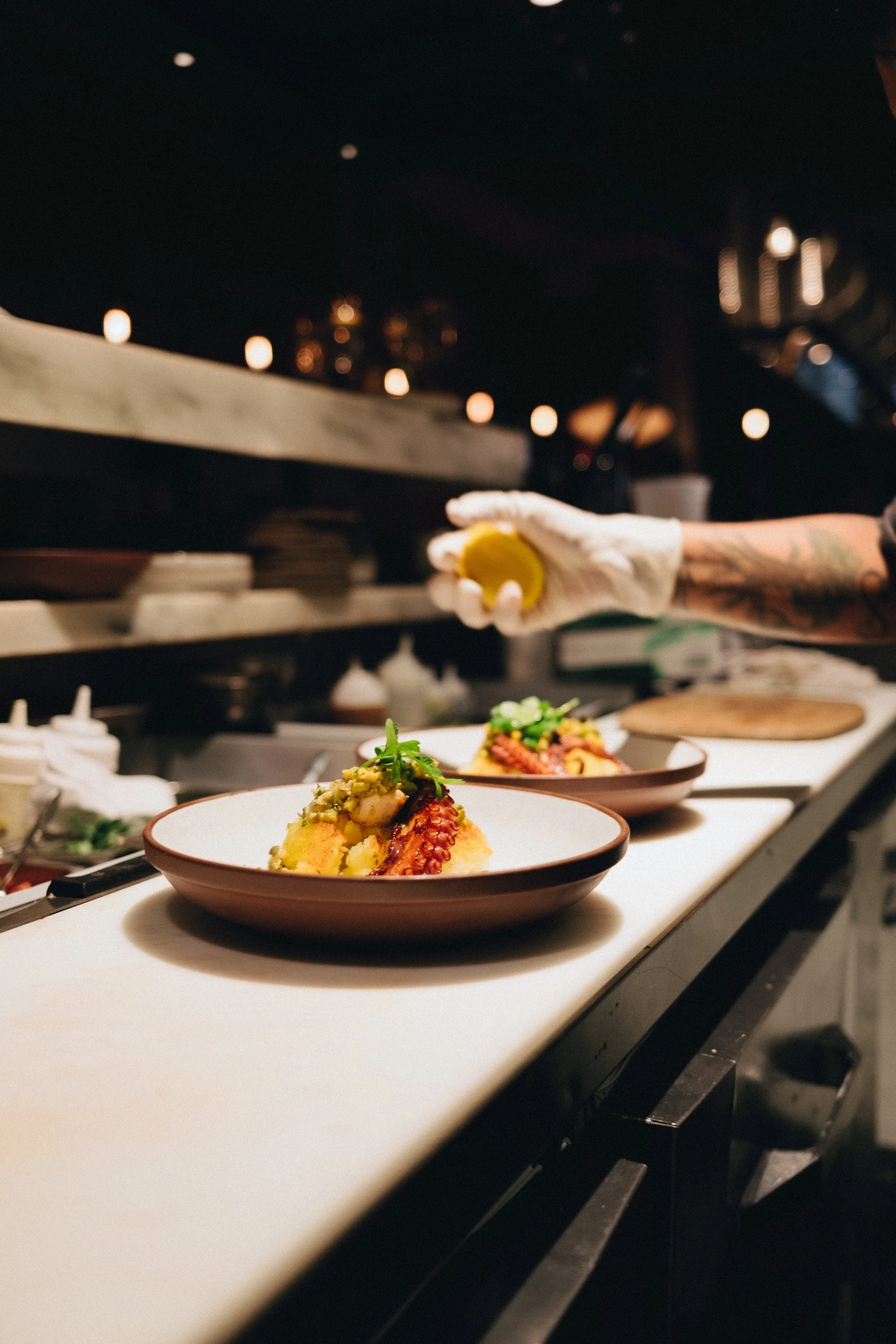 A chef wearing gloves and holding a lemon slice is preparing dishes in a professional kitchen with a dark background, warm bokeh lights, and plates of plated food being decorated.