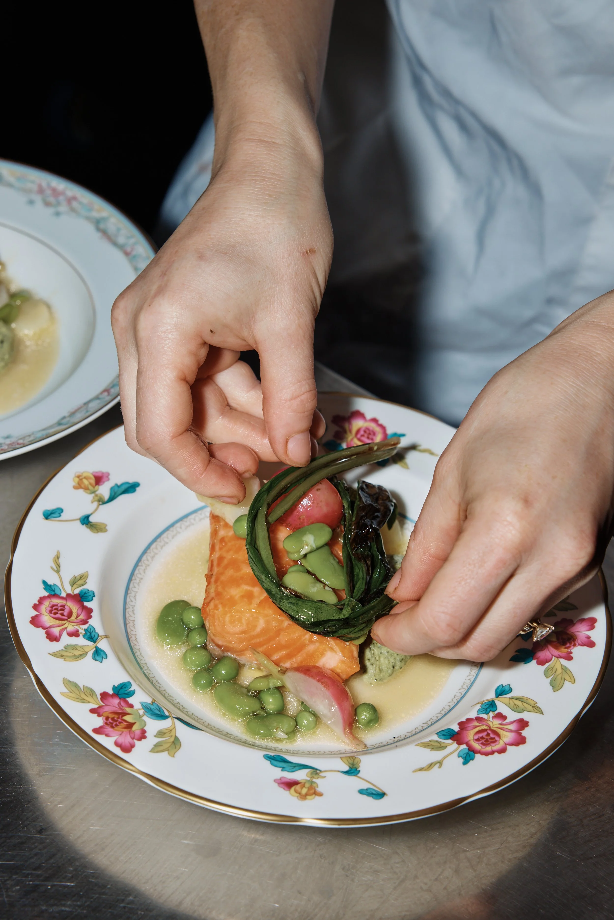 A person garnishing a plated dish with green beans, salmon, and vegetables on a floral-patterned plate.