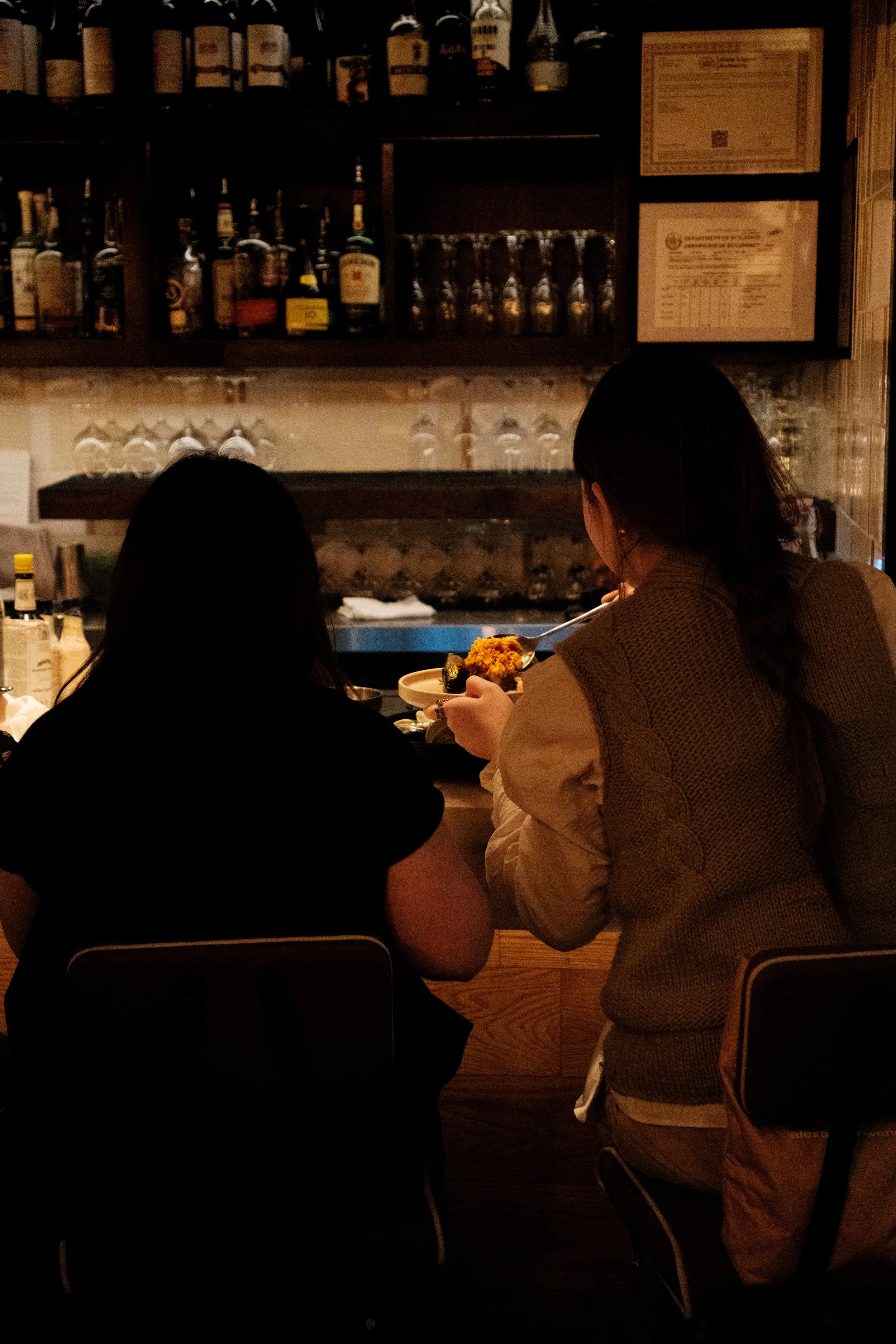 Two women sitting at a bar, one with dark hair and the other with short dark hair, having a conversation. The bar has bottles and glassware on shelves in the background.