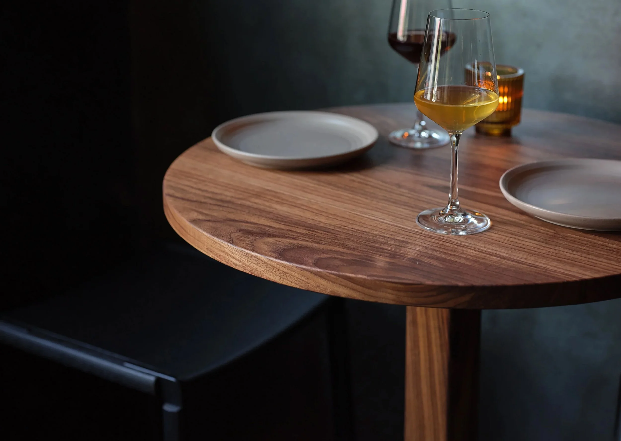 A wooden round table with two empty white plates, two glasses of wine, and one glass of beer, set against a dark background.