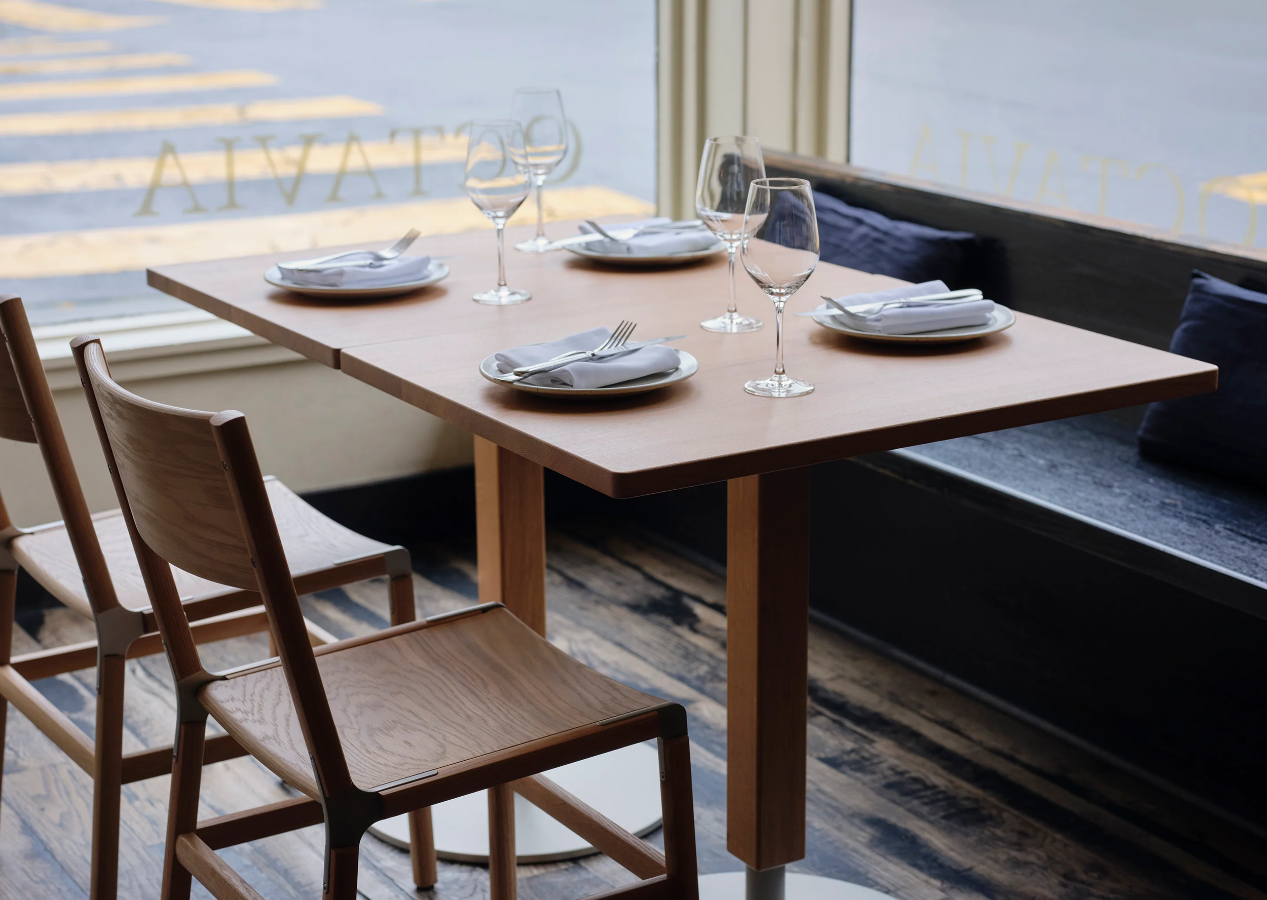 Empty restaurant table set with white napkins, plates, wine glasses, and silverware in front of a large glass window showing a street view.
