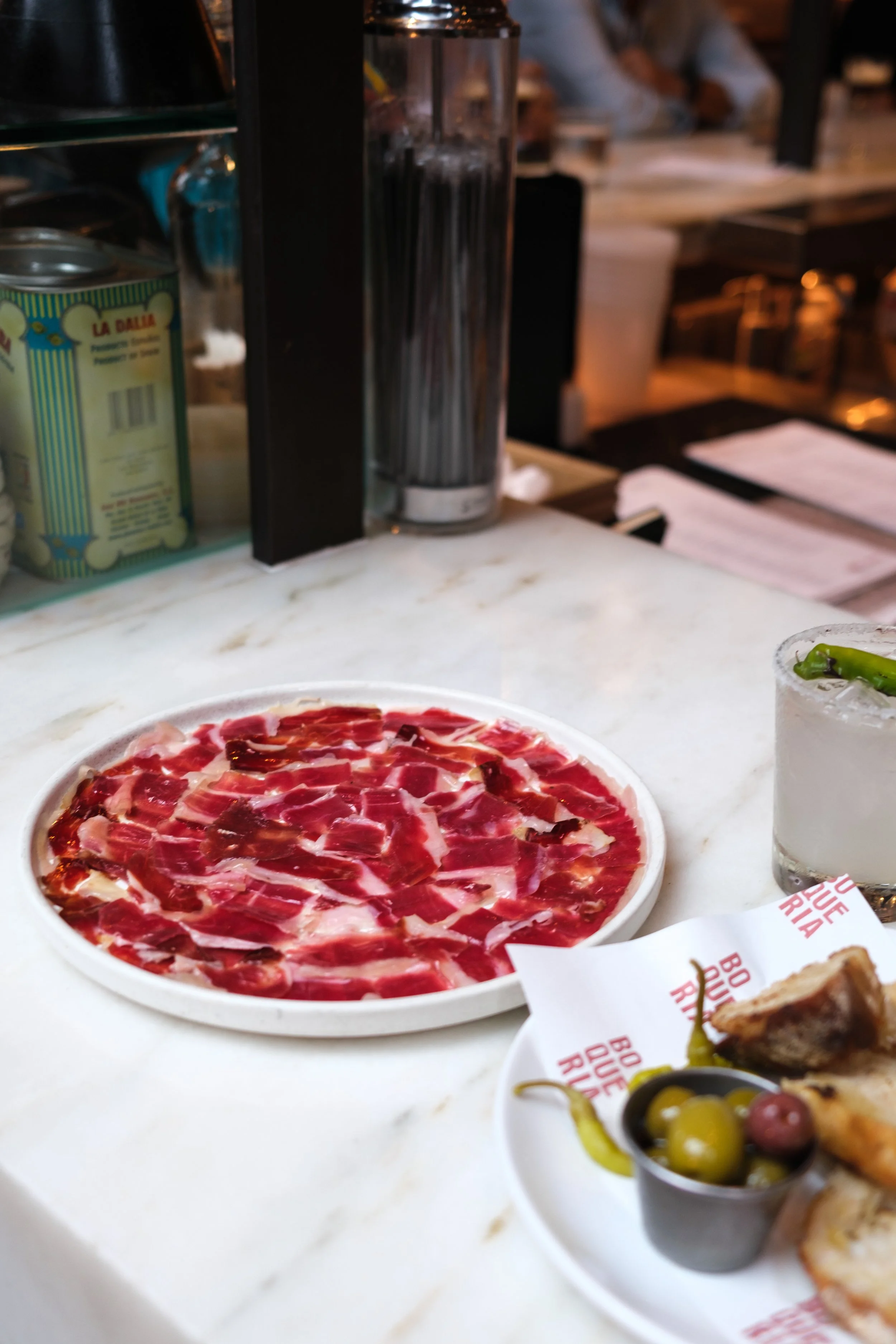 A plate of thinly sliced cured meat, a small cup of mixed olives, and a glass of what appears to be a beverage with ice and possibly herbs, on a white marble table at a restaurant.