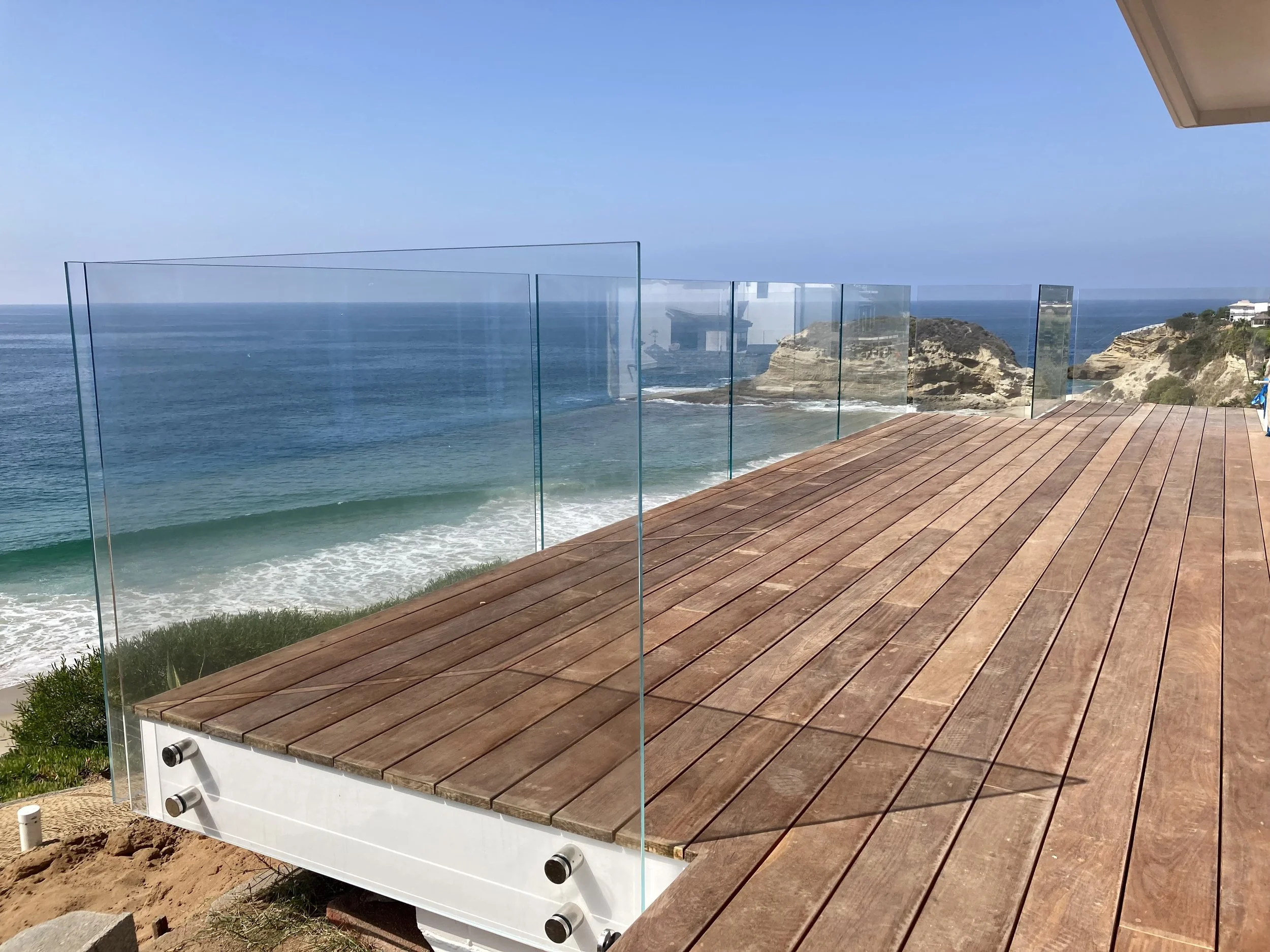 A wooden deck overlooking a large body of water with cliffs and houses in the distance. A glass railing surrounds the deck.
