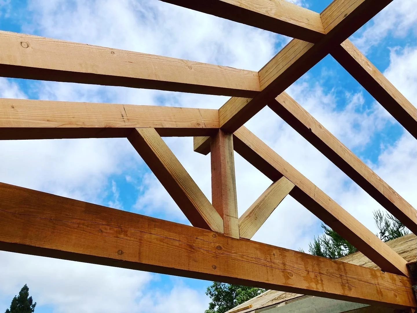 Wooden roof framework under blue sky with clouds, with trees visible in the background.