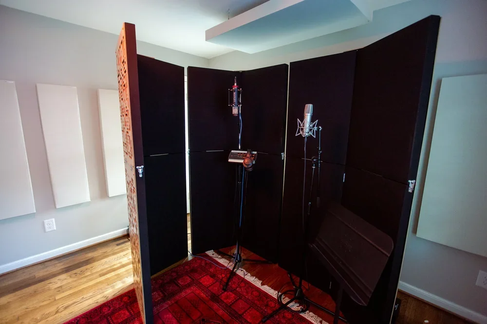 A recording studio with black soundproof panels, two microphones on stands, and a red carpet on a wooden floor.
