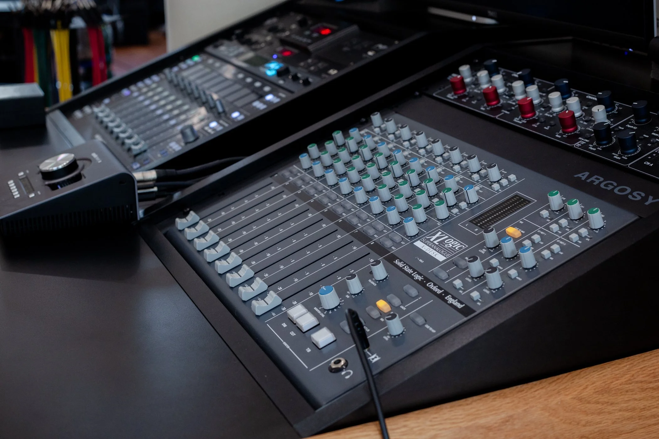 Audio mixing console with knobs and sliders on a desk with a computer monitor in the background.