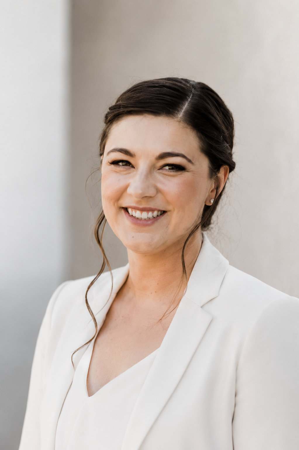 A woman with dark brown hair styled in loose waves, wearing a white blazer and a white top, smiling against a neutral background.
