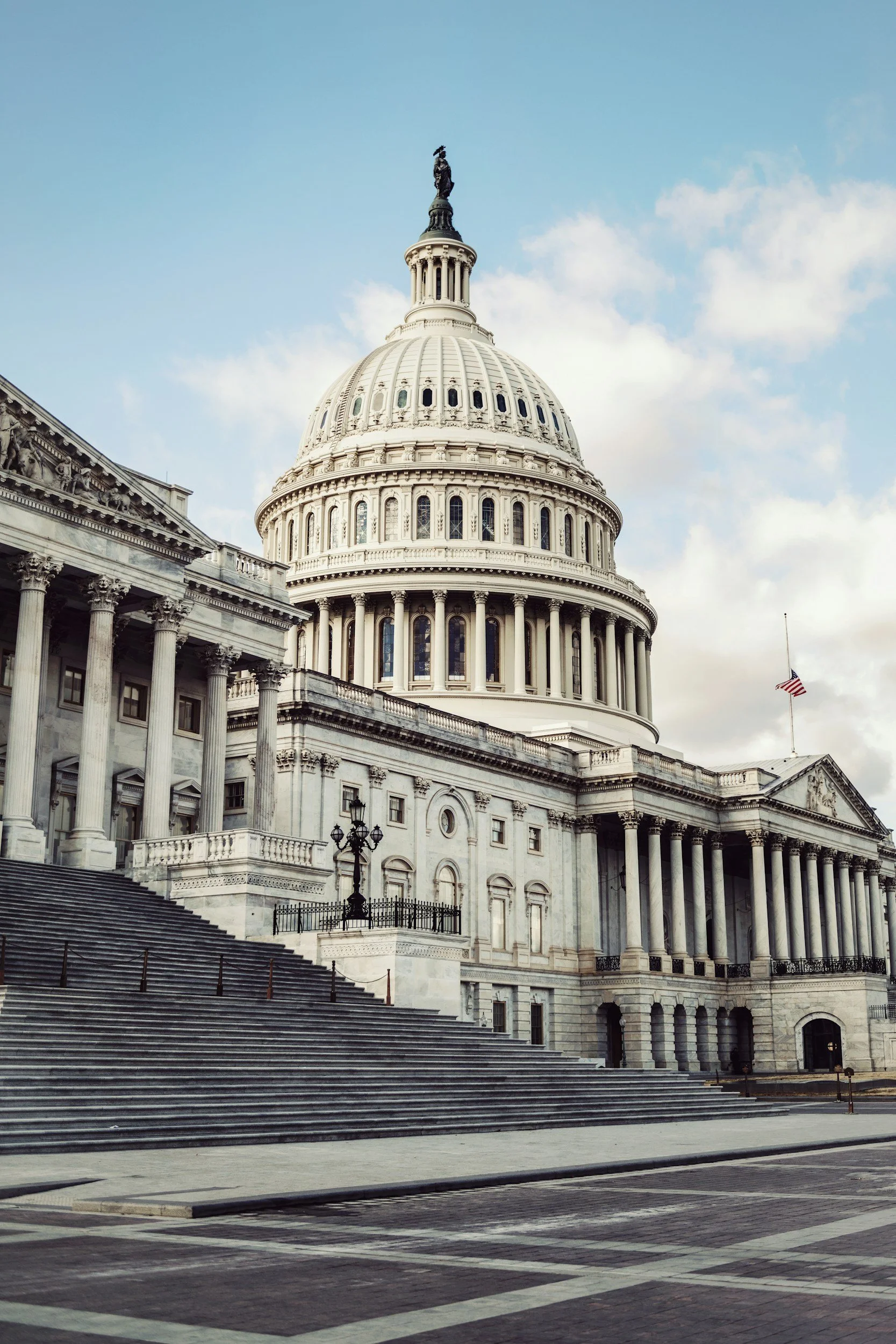 The United States Capitol building in Washington, D.C., with its iconic dome and neoclassical architecture, featuring wide steps leading up to the entrance and a flag flying in the foreground.