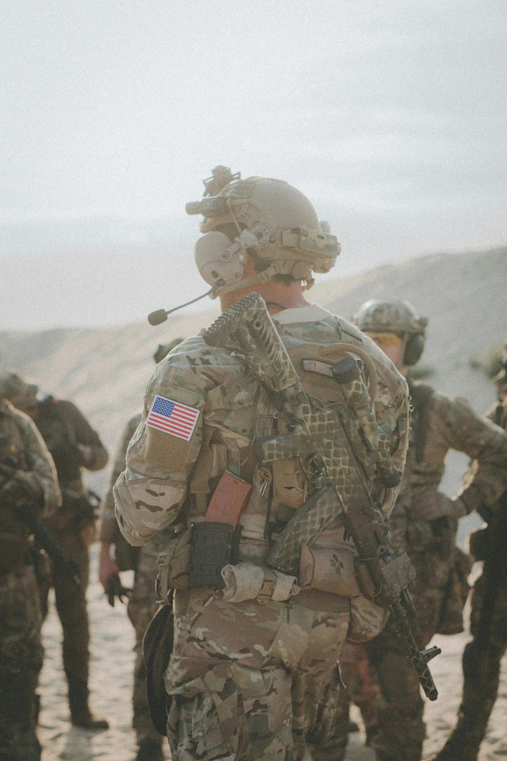A group of soldiers in desert camouflage uniforms standing outdoors with helmets and tactical gear, some with American flags on their sleeves, in a dusty landscape with hills in the background.