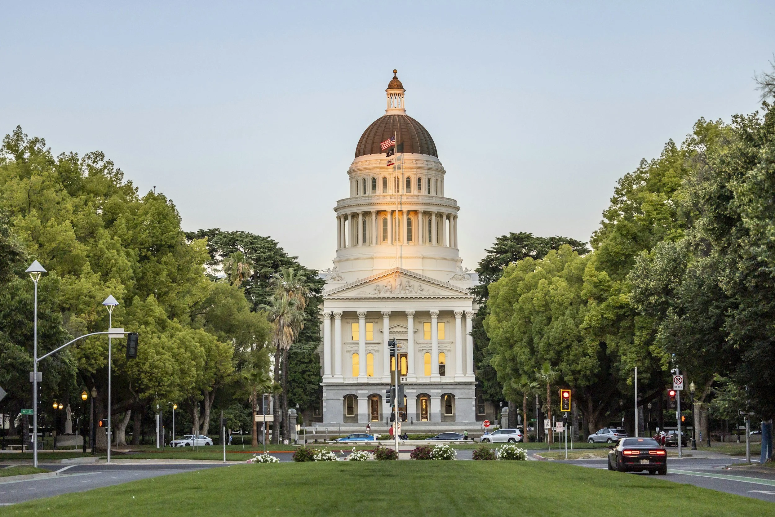 The California State Capitol building in Sacramento, surrounded by trees, cars, and street lights during daylight.