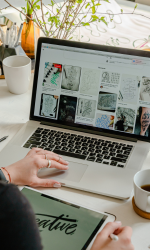 A person using a laptop to browse a website displaying digital artwork or illustrations, with a tablet in hand, on a white desk with coffee cups and a plant.