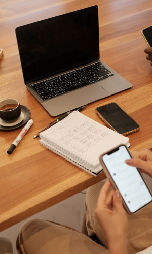 A wooden desk with a silver laptop, a black smartphone, a white notebook with handwritten notes, a white marker, a coffee cup on a saucer, and a person's hand holding a smartphone.
