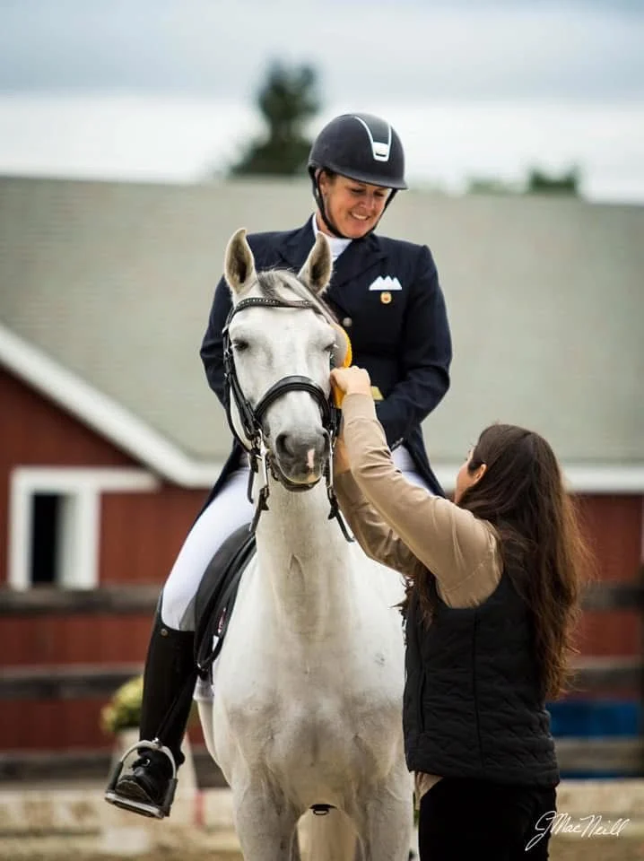 A woman wearing a riding helmet and formal riding attire is smiling while sitting on a white horse. Another woman, dressed in casual clothing, is standing next to the horse and adjusting the rider's helmet strap.