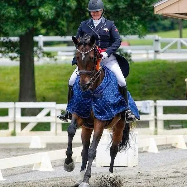 A woman riding a horse during a dressage competition or practice, with wooded background and white fences.