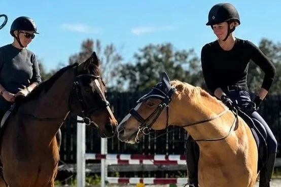 Two women riding horses and wearing helmets, exchanging a friendly look in an outdoor riding arena.