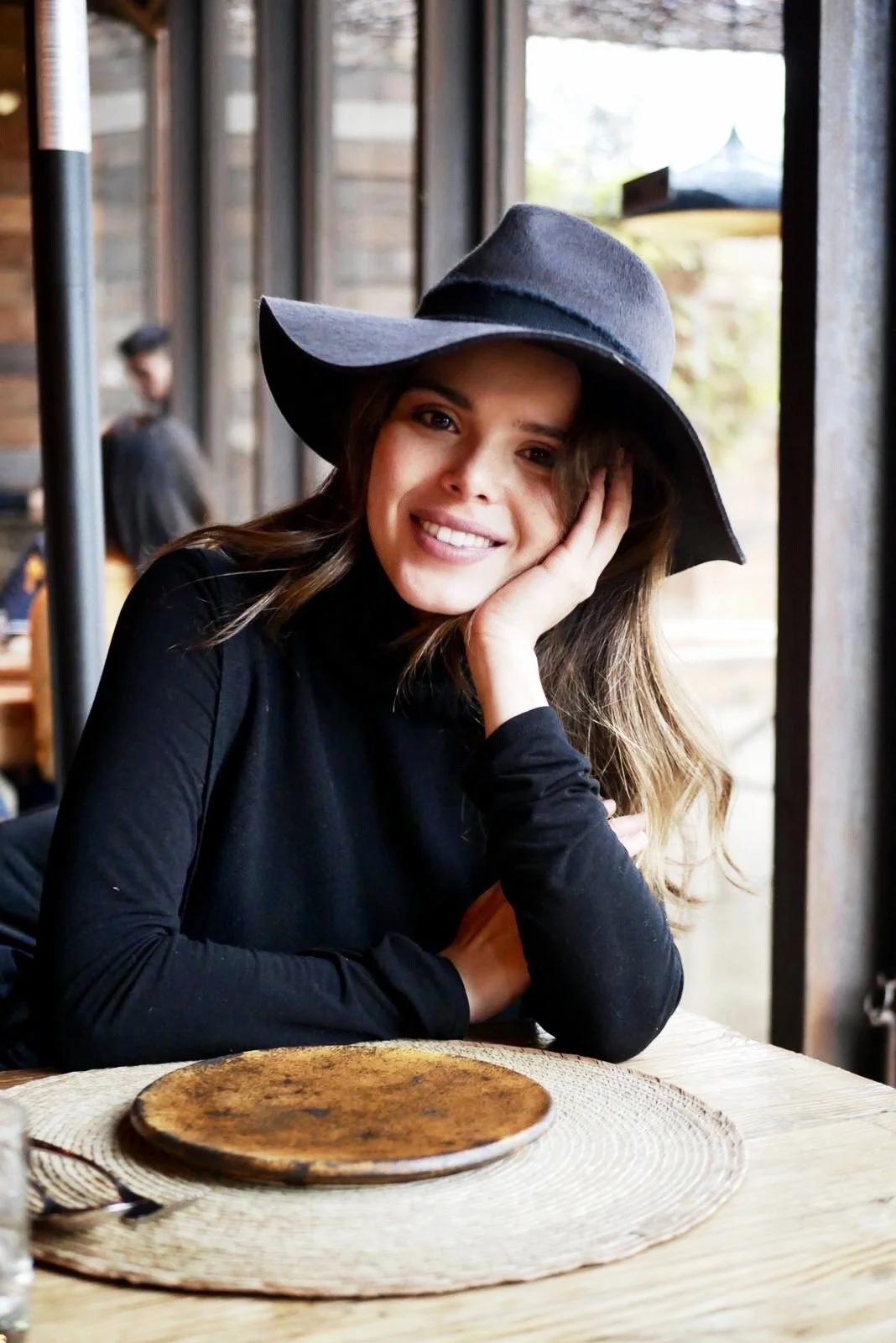 A woman wearing a black top and a large black hat sitting at a wooden table with a round wooden plate in front of her, smiling at the camera with her head resting on her hand.