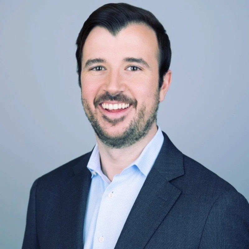 Professional headshot of a smiling man with dark hair, a beard, wearing a navy blazer and light blue shirt against a plain light gray background.