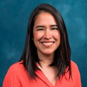 A woman with long dark hair smiling, wearing a coral top, against a teal background.
