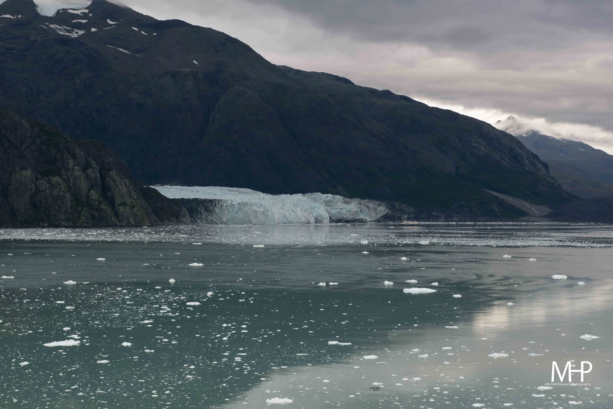 A view of a glacier calving into a body of water with floating ice chunks, mountains in the background, and overcast sky.