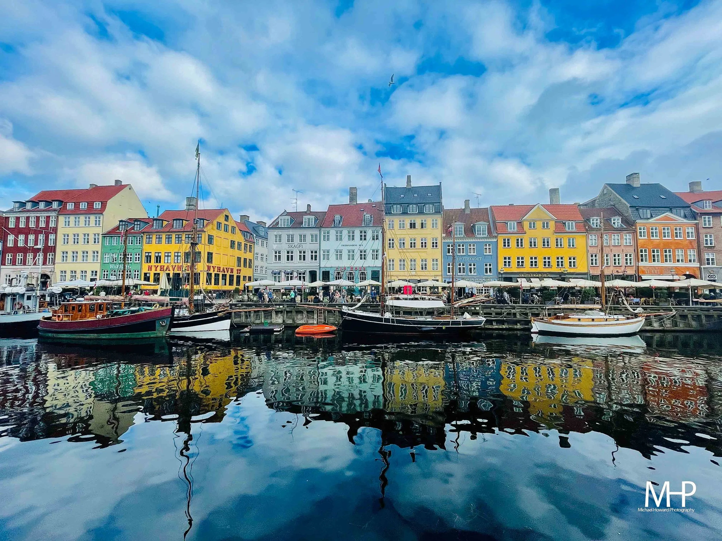Colorful buildings line a waterfront in this cityscape, with boats docked at the marina and a blue sky with scattered clouds overhead.