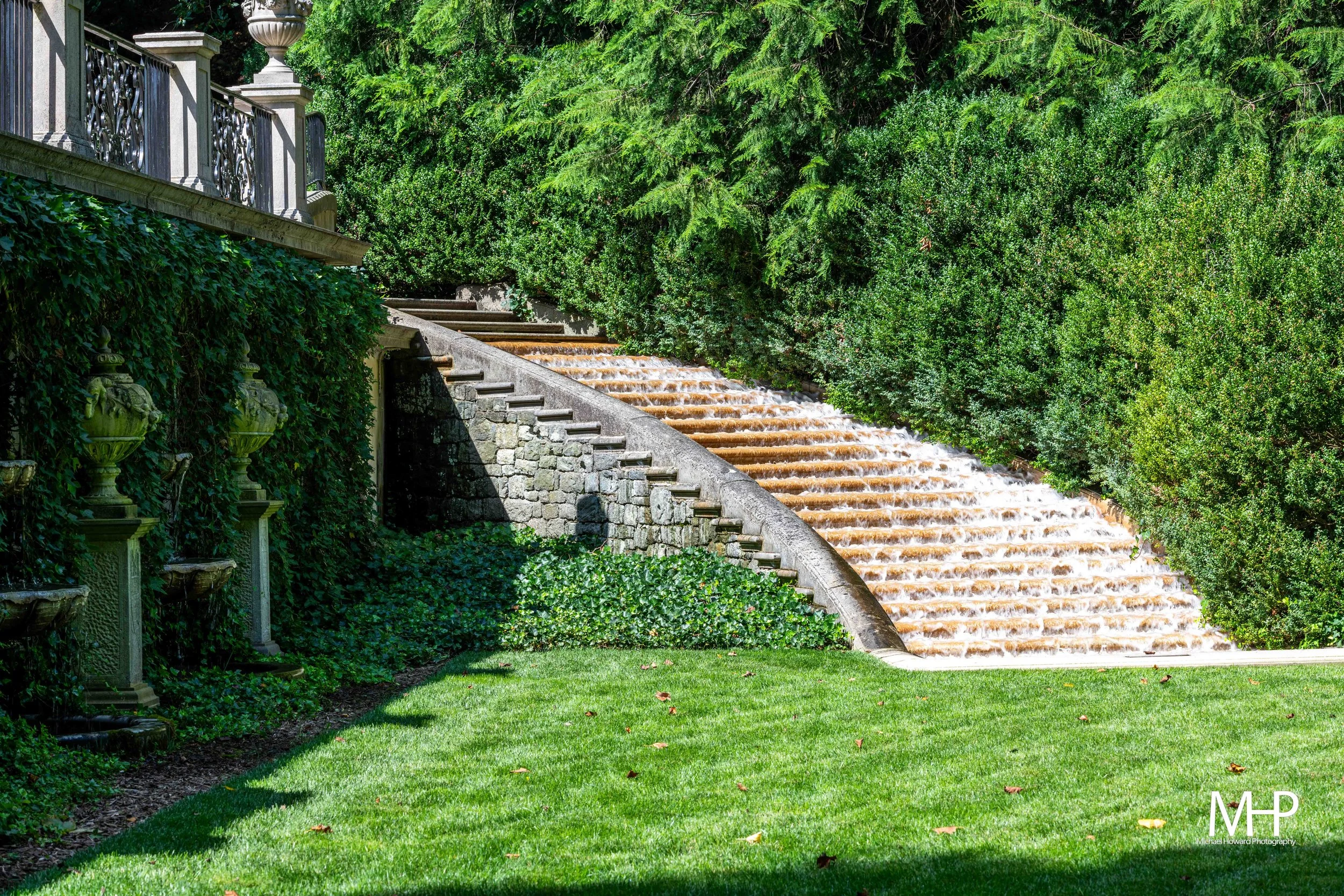 A staircase with water flowing down, surrounded by lush green trees and bushes, with a stone railing and decorative urns on the left edge.