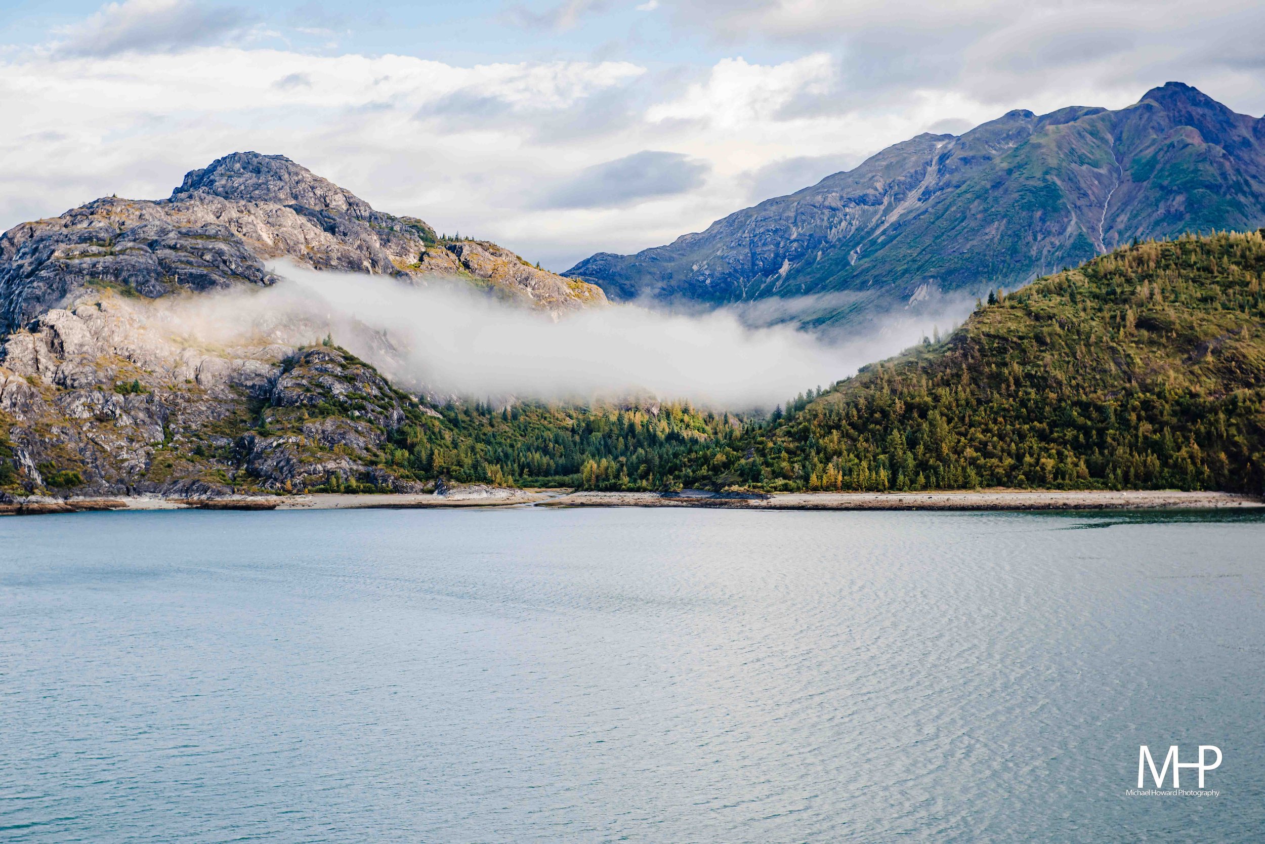 Mountain landscape with rocky peaks, green forested slopes, fog near the mountain base, and a calm body of water in the foreground.