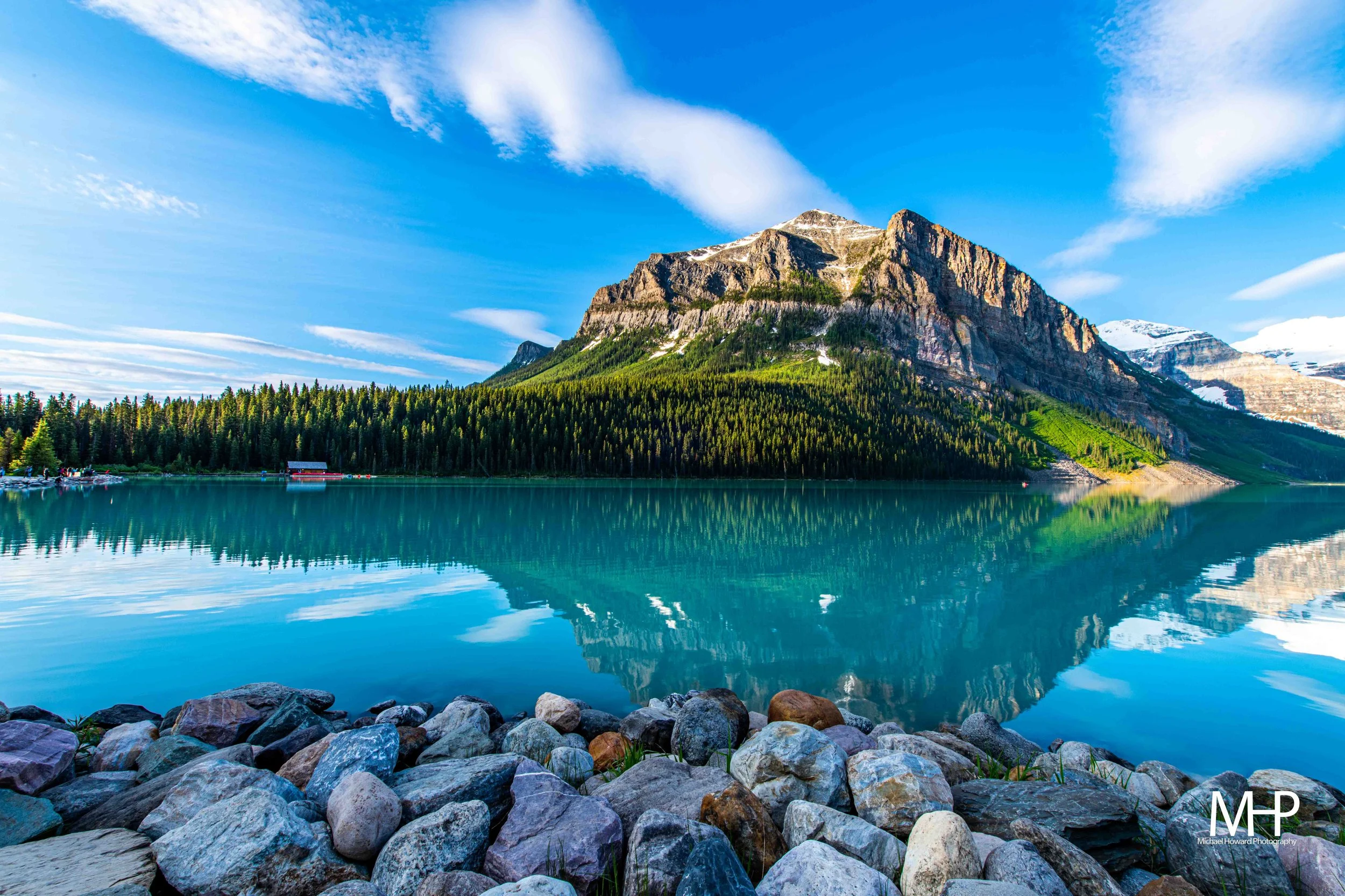 A scenic view of a mountain with a snow-capped peak reflected in a calm, clear lake, with a rocky shoreline in the foreground and a forested hillside beneath the mountain.
