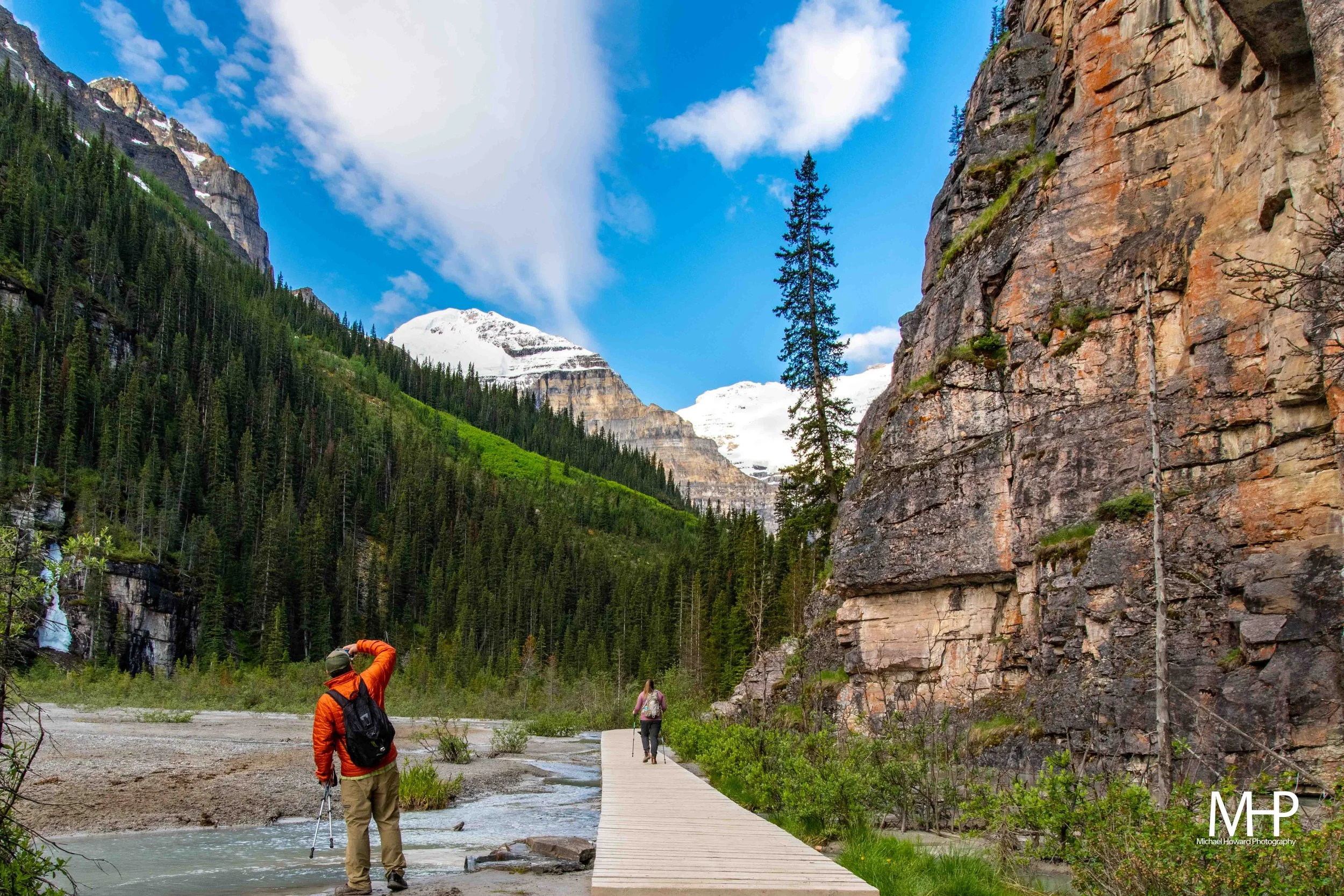 People hiking in a mountainous area with cliffs, tall pine trees, and snow-capped peaks in the background. A wooden trail runs along a small creek on a cloudy day.
