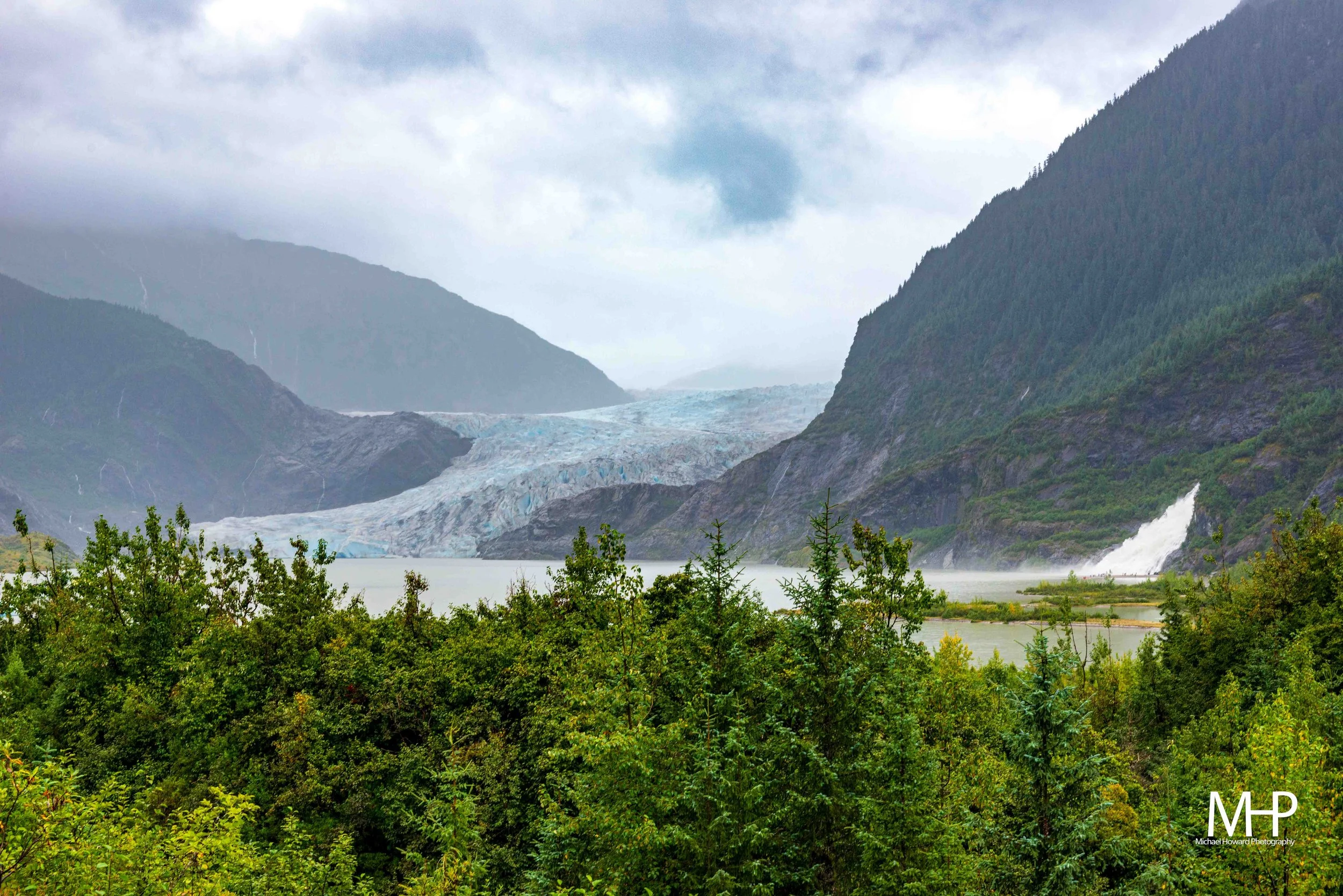 A scenic landscape featuring a glacier nestled between green, forested mountains under a cloudy sky.