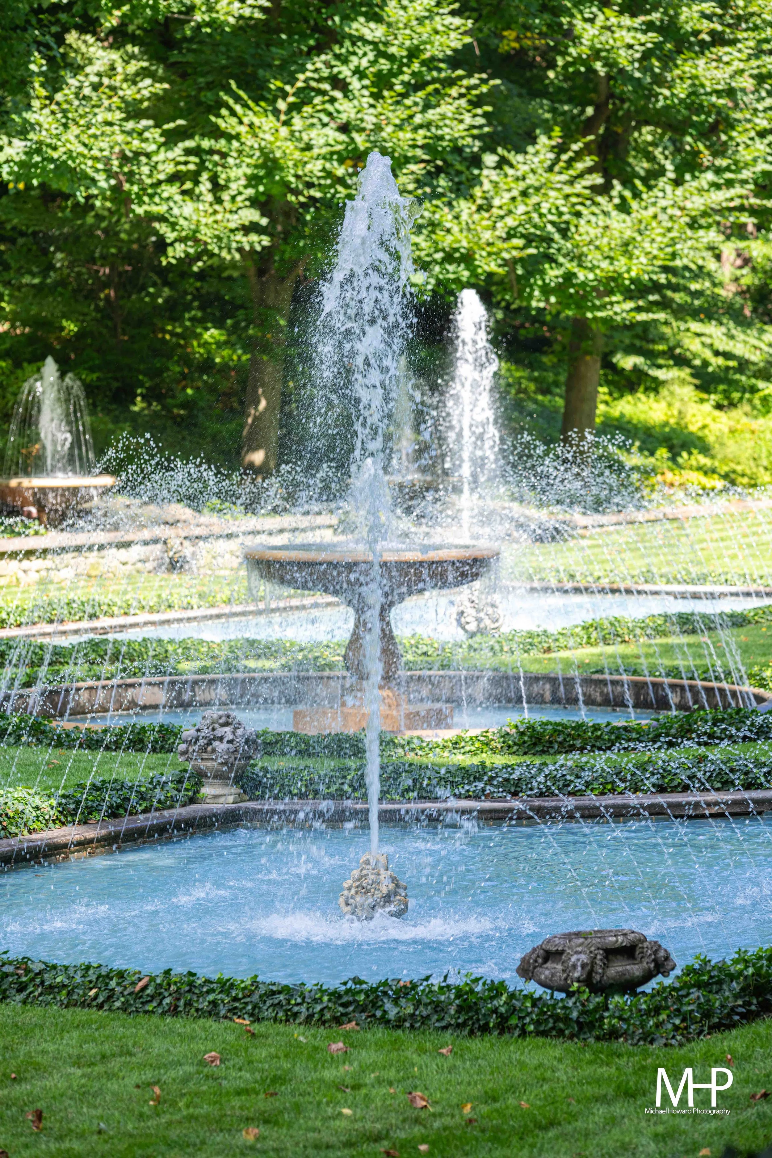 A fountain with water spouting from multiple jets into a round basin, surrounded by lush greenery and trees.