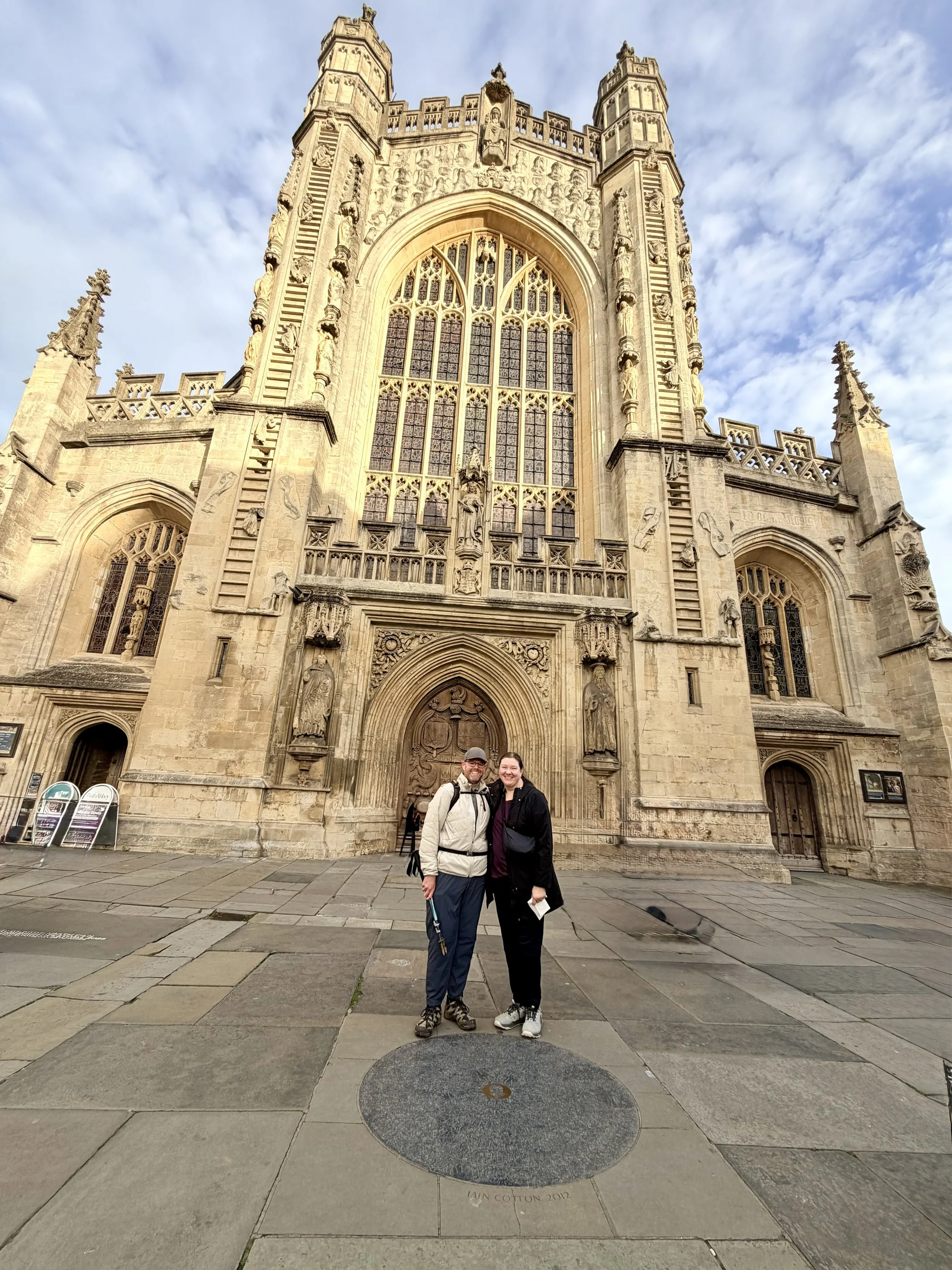 Two people standing together in front of an ornate gothic-style church, smiling at the camera. The church has intricate stone carvings, large stained glass windows, and tall spires reaching into the sky. The ground is paved with stone tiles.