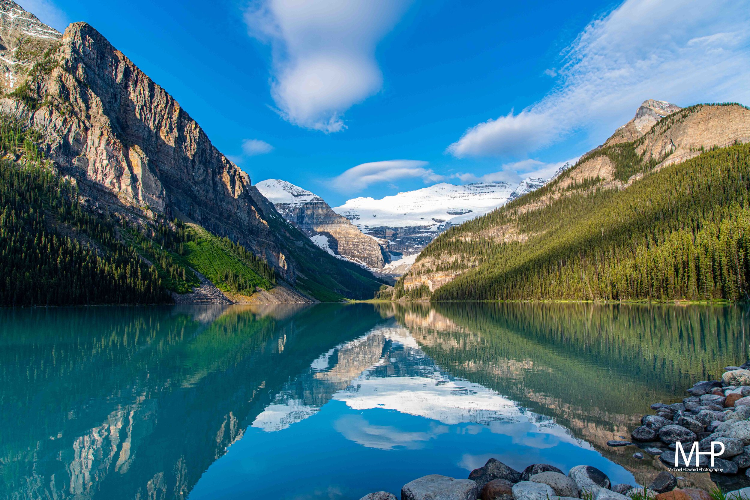 A mountain landscape with a lake in the foreground, surrounded by forested slopes and snow-capped peaks under a partly cloudy sky.