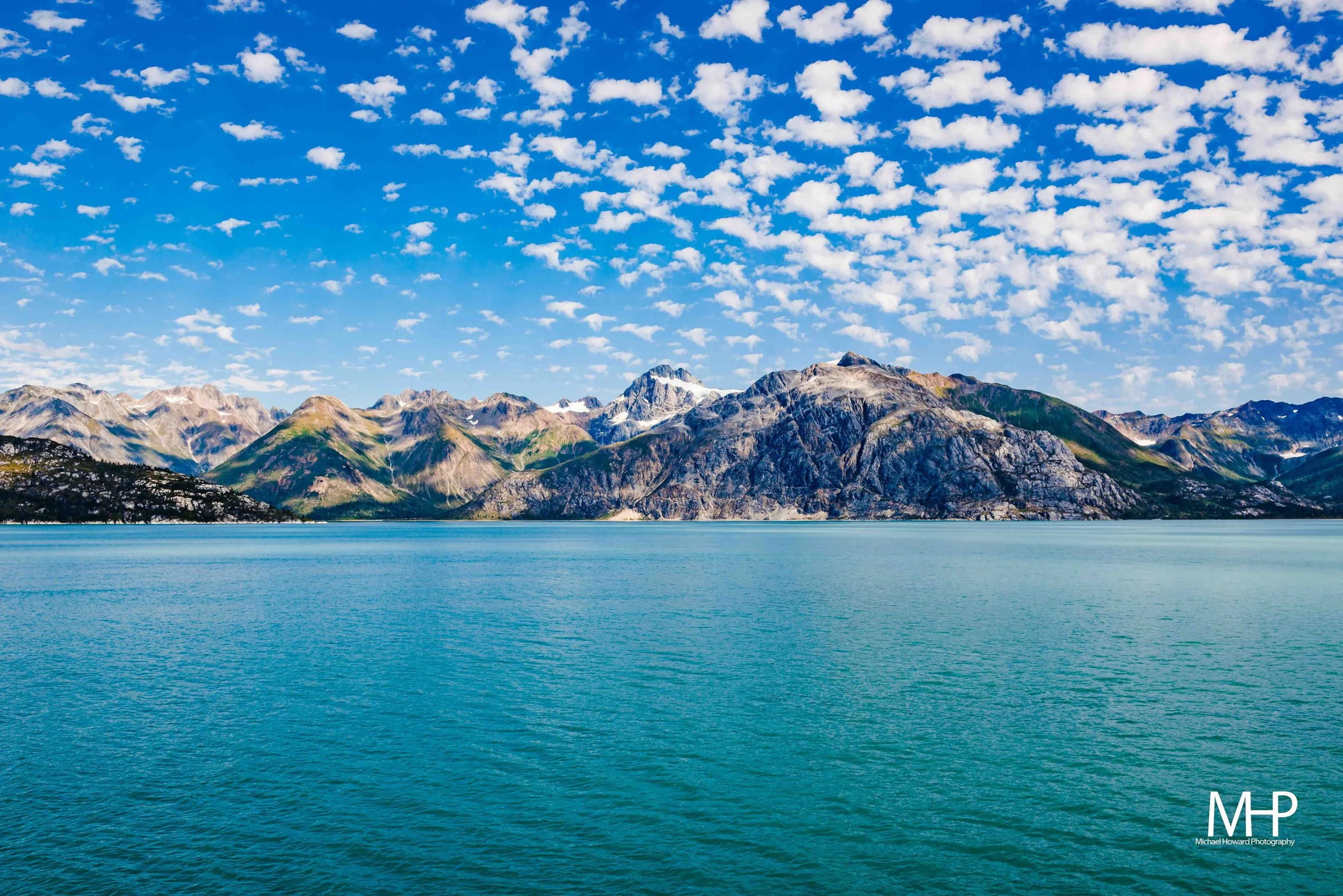 Scenic view of a mountain range with rugged peaks and patches of snow, seen across a large body of turquoise water, with a bright blue sky filled with scattered white clouds.