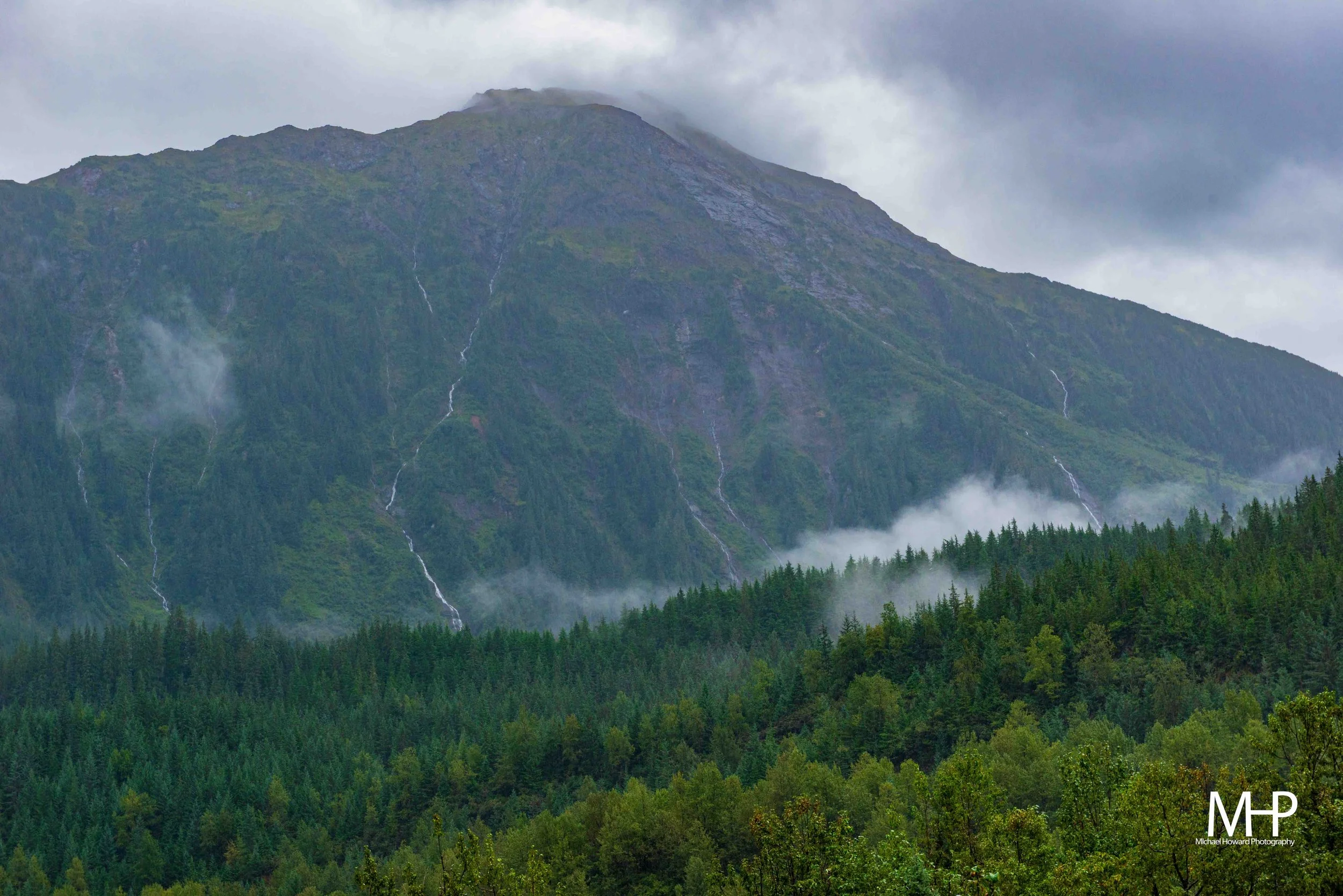 A mountain with patches of greenery and multiple waterfalls cascading down its slopes, surrounded by a dense forest and cloudy skies.