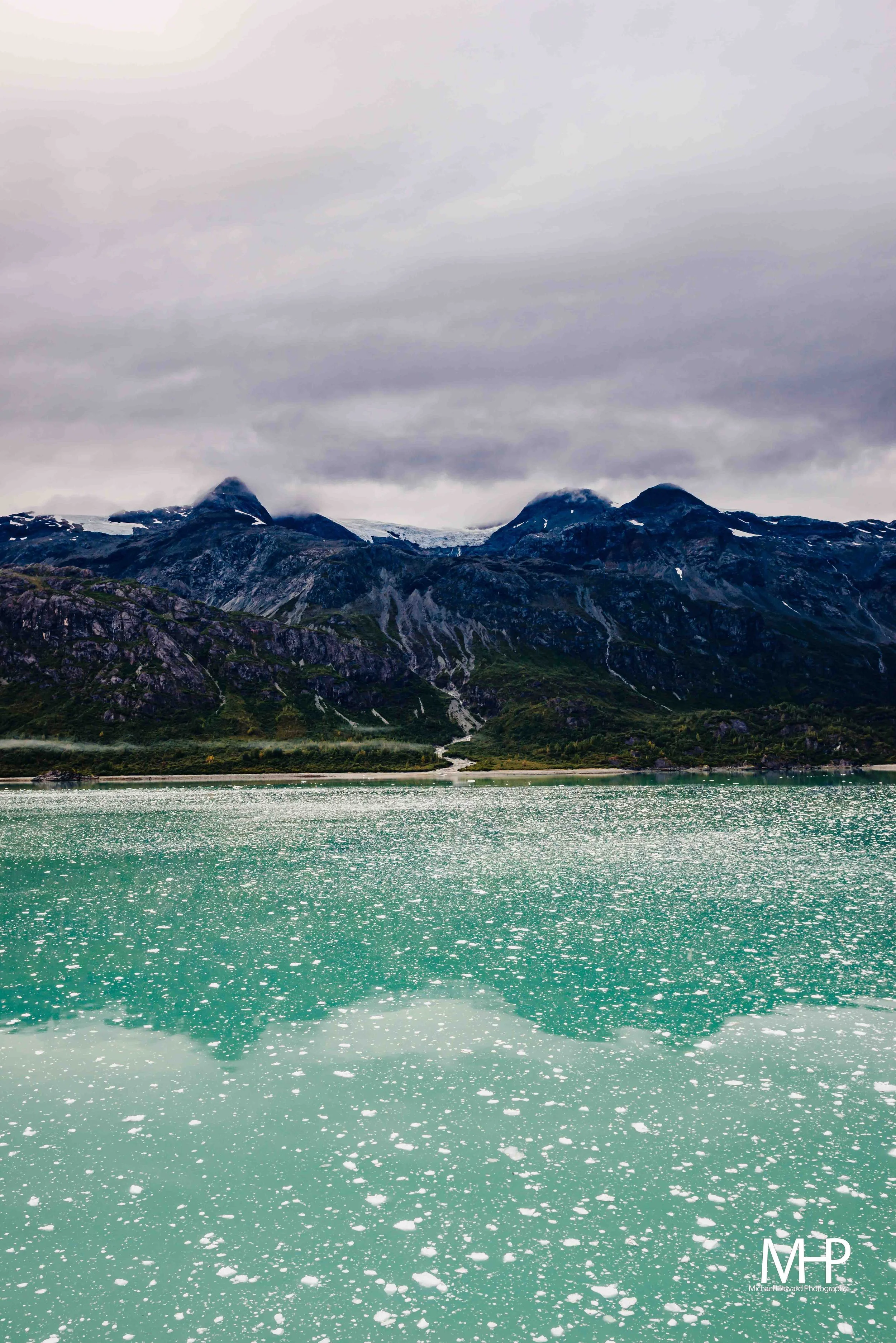 A mountain range with rocky peaks and some snow patches under a cloudy sky, reflected on a body of water with floating ice.