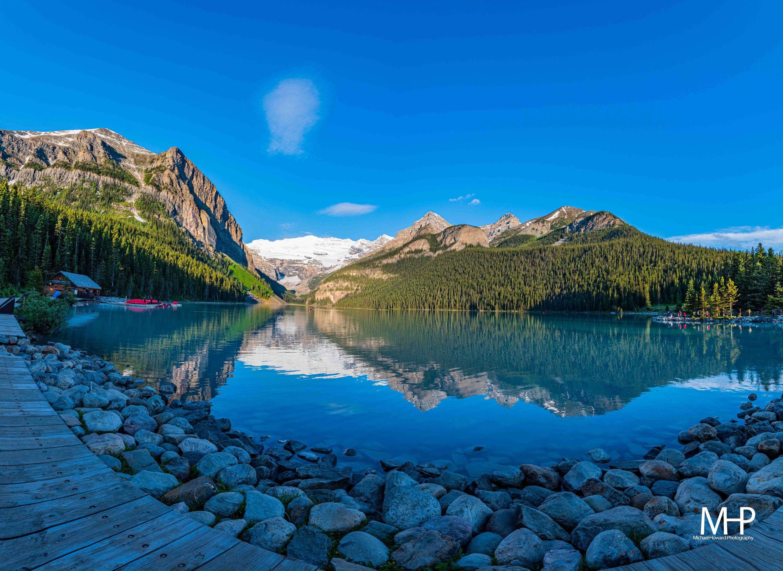 A scenic mountain landscape featuring a clear lake with a wooden shoreline, lush green pine forests, towering mountains with some snow patches, and a sky with a few scattered clouds.