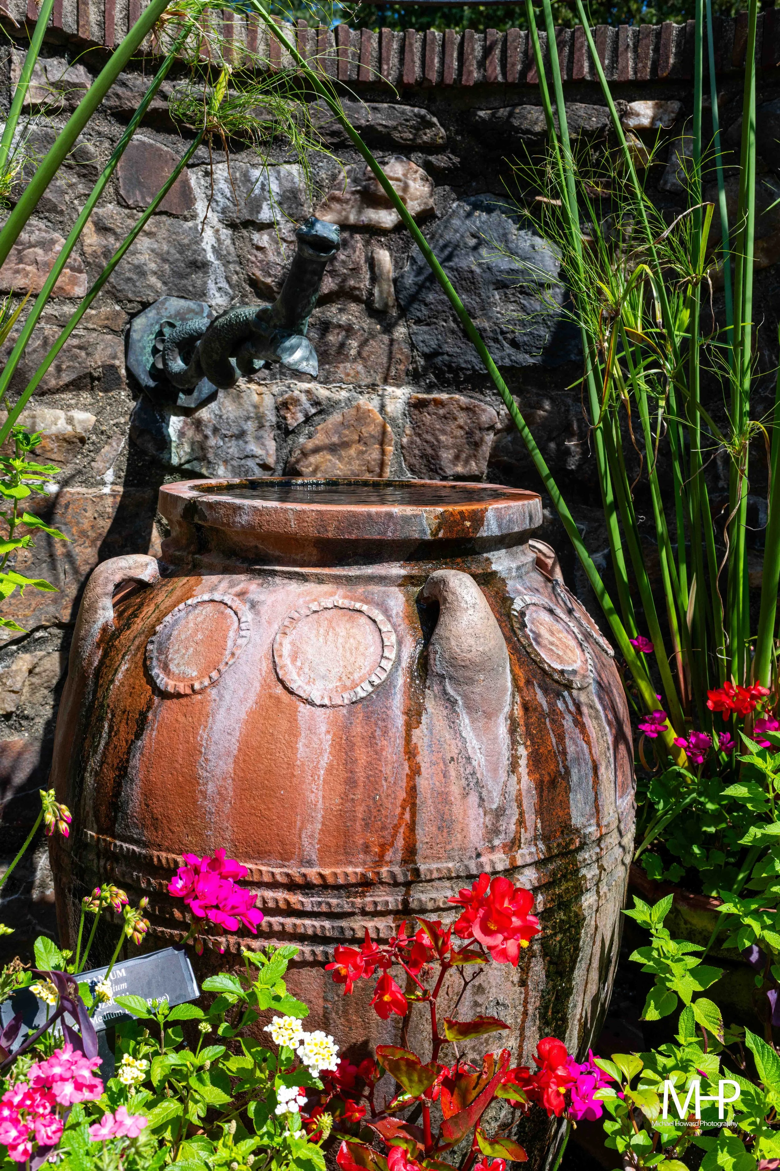 A decorative stone water fountain set against a stone wall, surrounded by vibrant pink, red, white, and purple flowers and green plants in a garden.