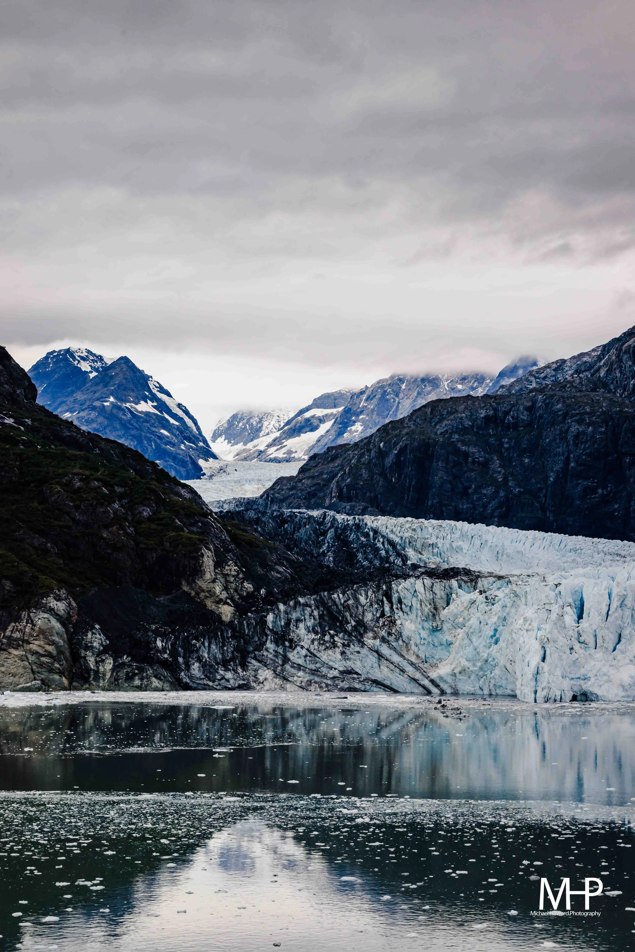 A scenic landscape of a snowy mountain range with glaciers, dark rocky cliffs, and a reflective body of water in the foreground under a cloudy sky.
