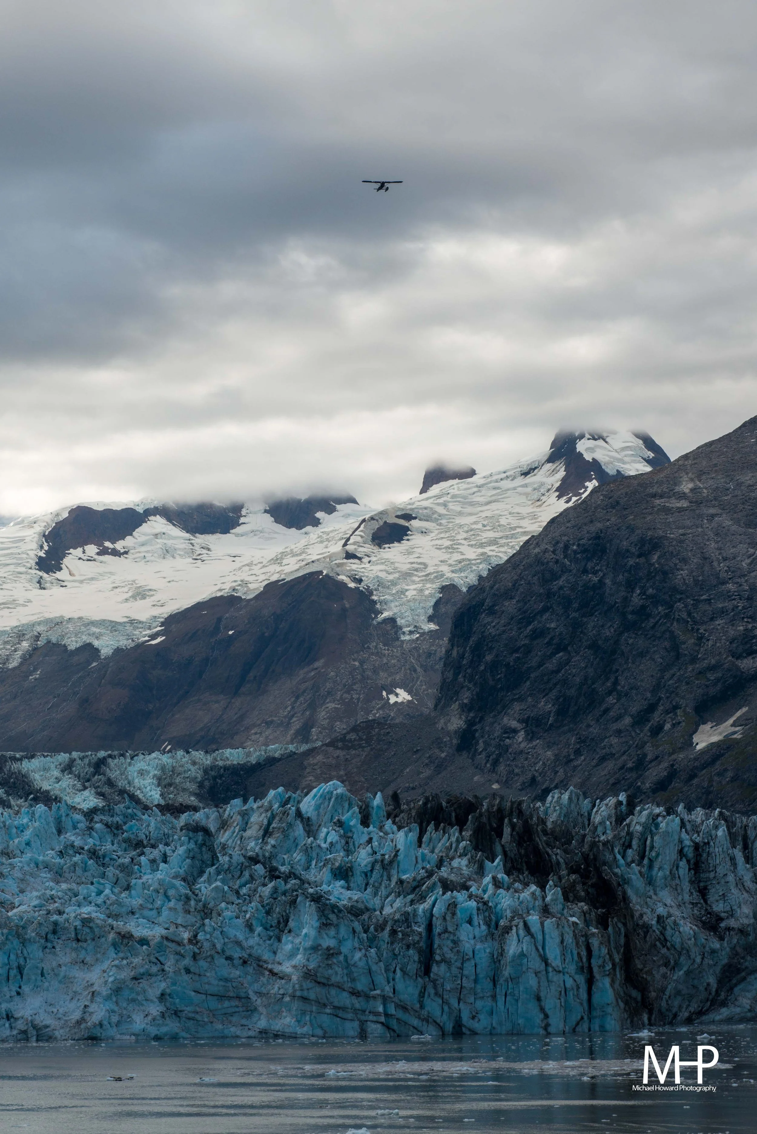 A glacier with blue ice extending into water, with snow-covered mountains and a dog flying a small airplane in the cloudy sky above.