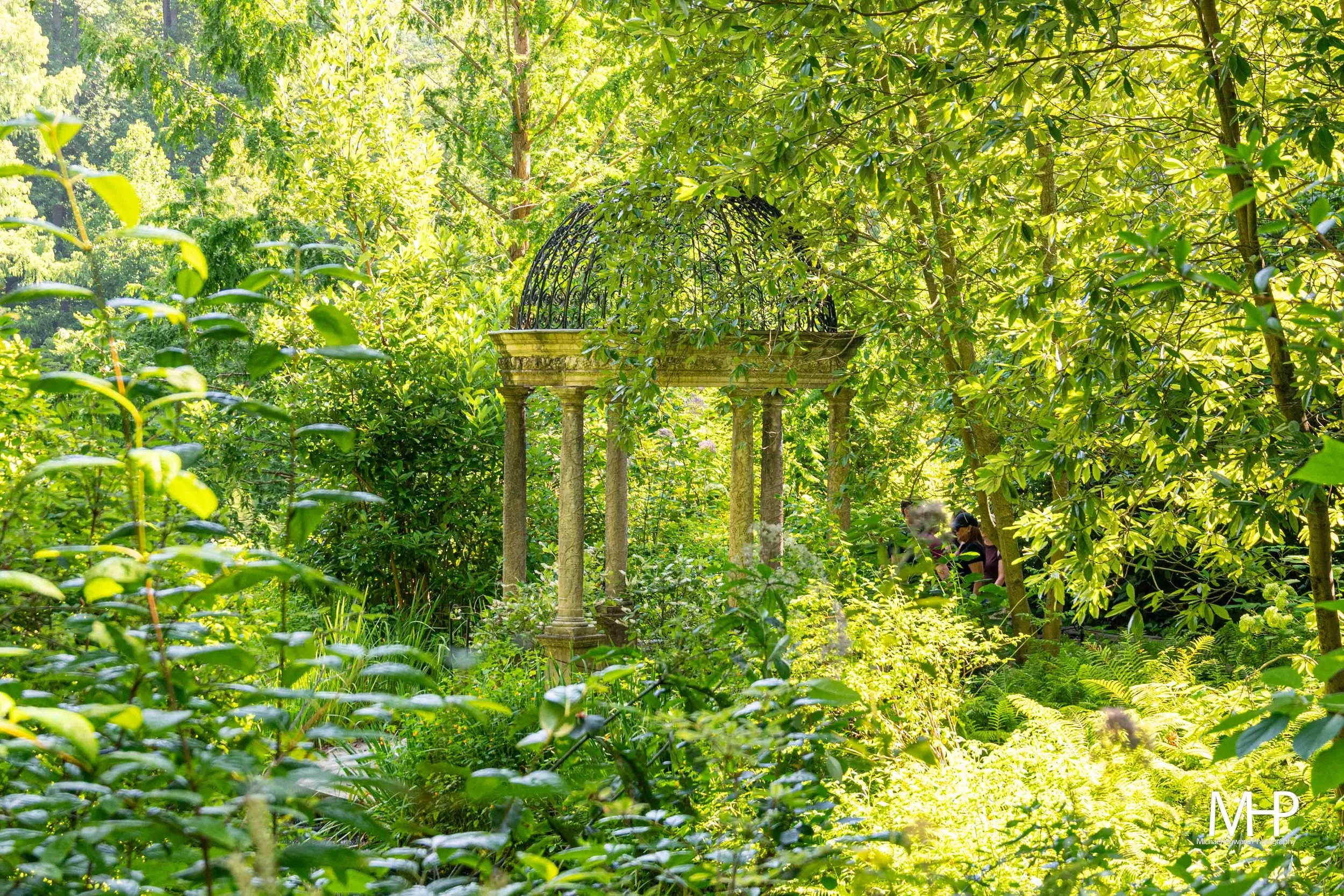 A lush, green garden with tall trees and dense foliage, featuring a stone pedestal with a decorative black metal cage or top on it, surrounded by sunlight filtering through the leaves. There are people partially visible on the right side of the image