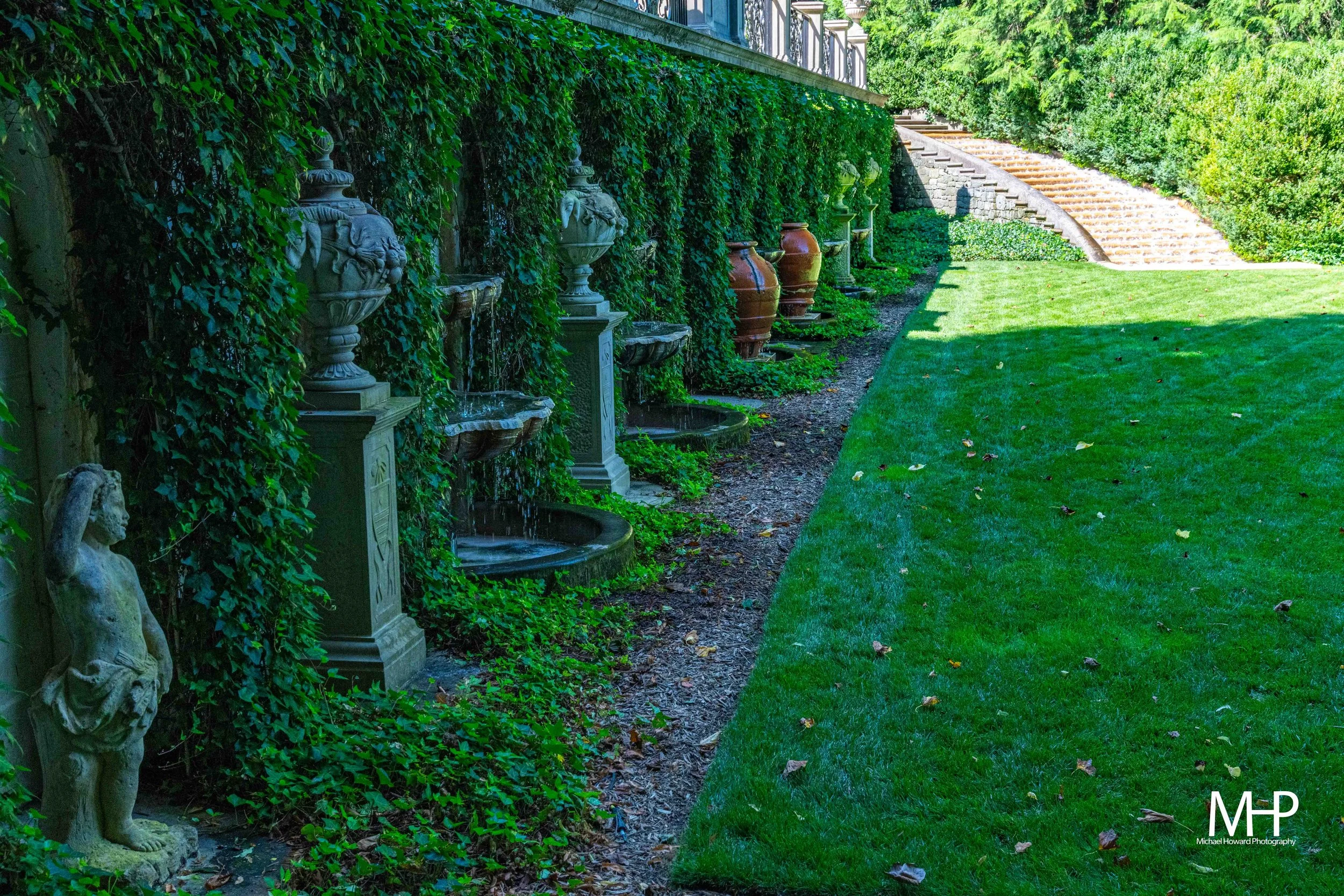 A garden with a wall covered in green ivy, featuring decorative water fountains and urns along the base, a staircase to the right, well-manicured grass, and a bright, sunny atmosphere.