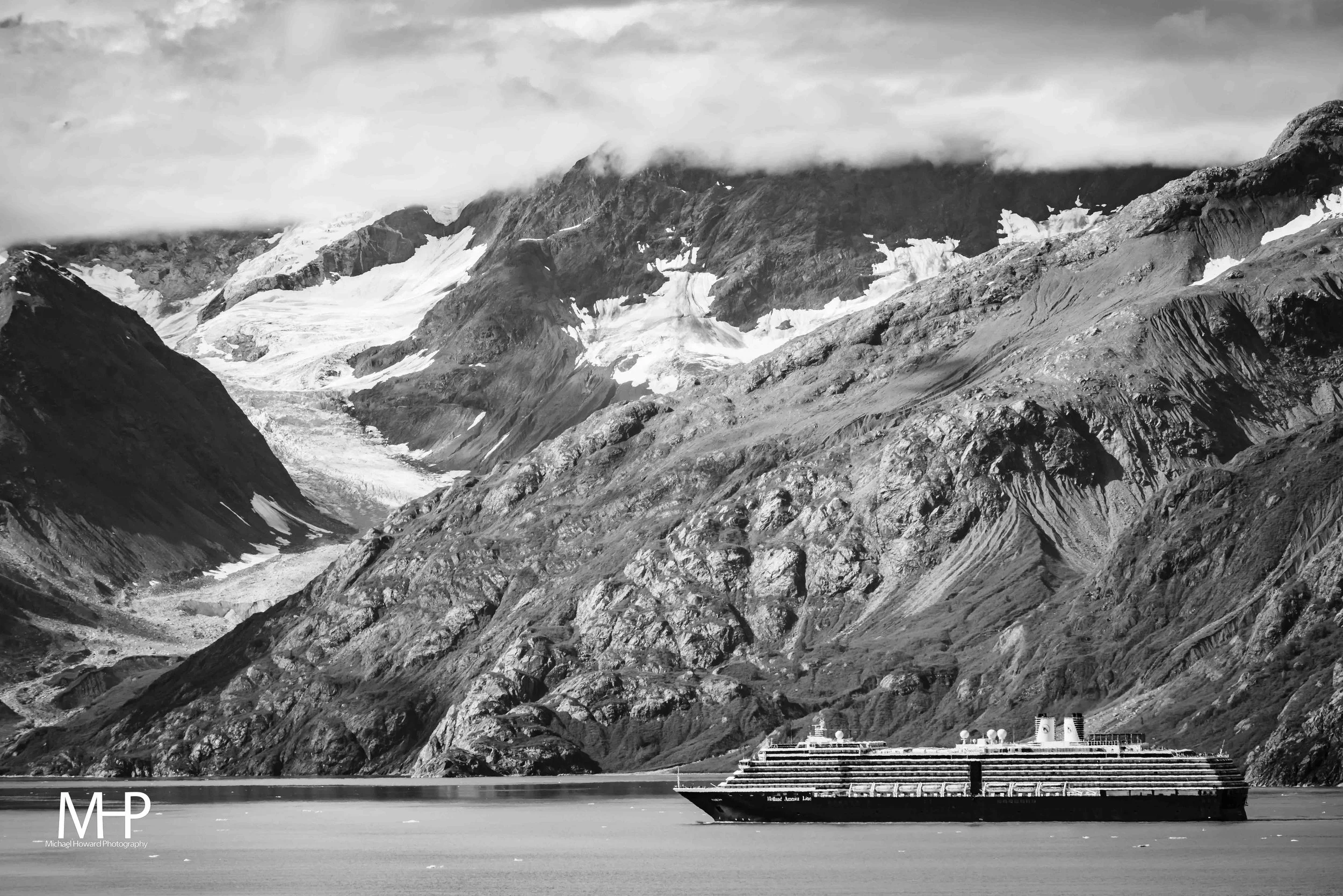 A black and white photograph of a cruise ship sailing in a body of water, with rugged mountains and glaciers in the background, partly covered by clouds.