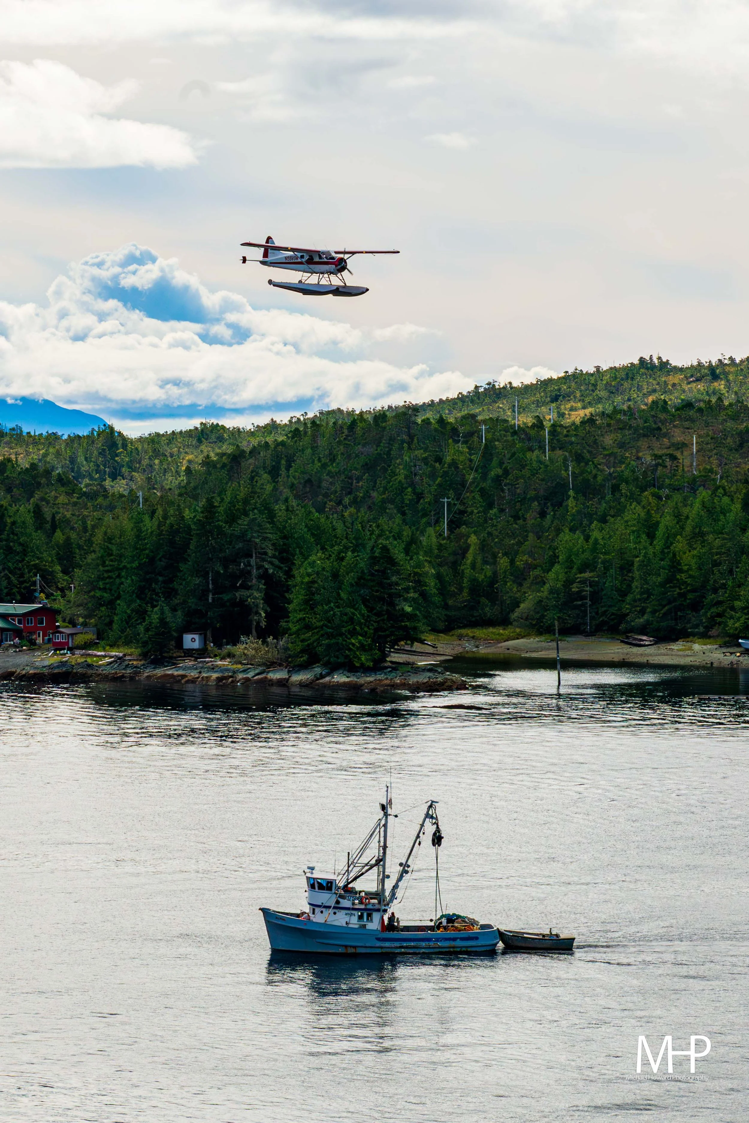 A peaceful scene of a fishing boat on calm water with a lush green hillside in the background. A small airplane is flying in the cloudy sky above.