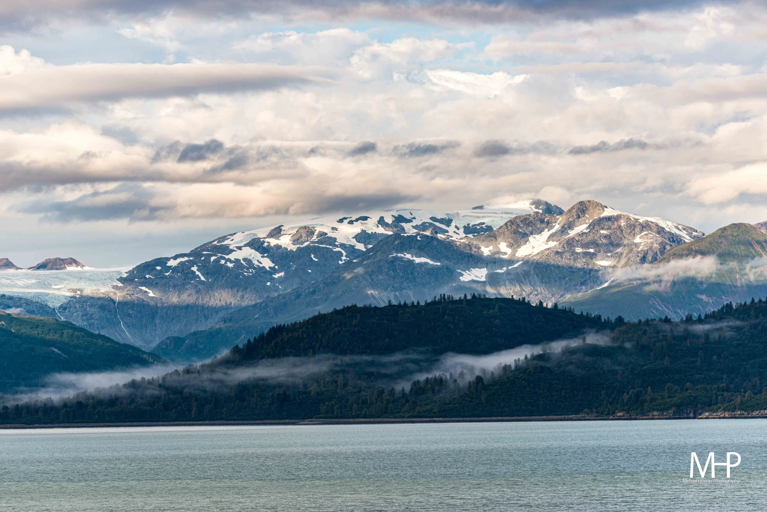 Scenic view of snow-capped mountains with glaciers, green forested slopes, and a body of water in the foreground under a cloudy sky.