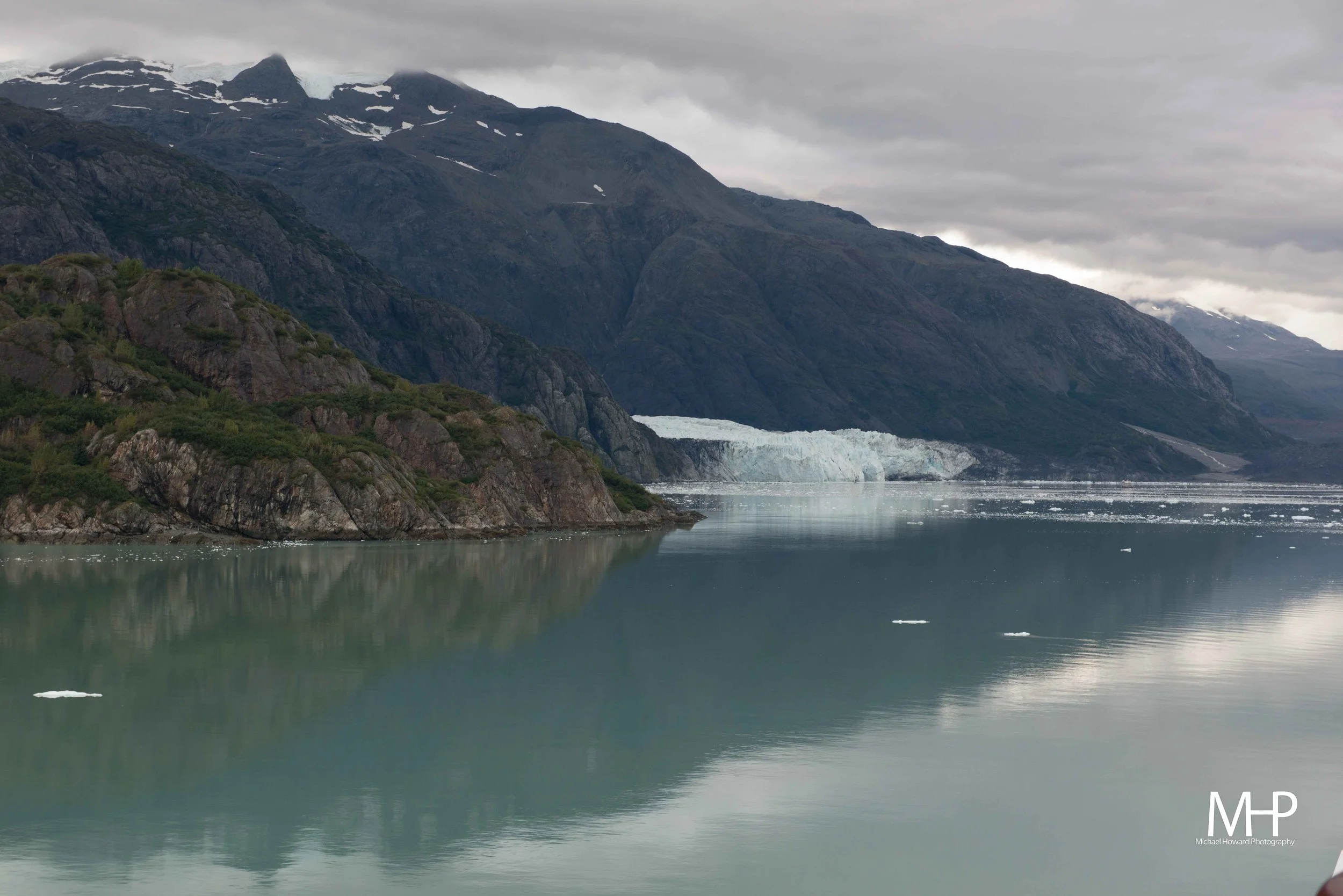A scenic view of a mountain range with snow patches and glaciers, reflected in a calm body of water under a cloudy sky.